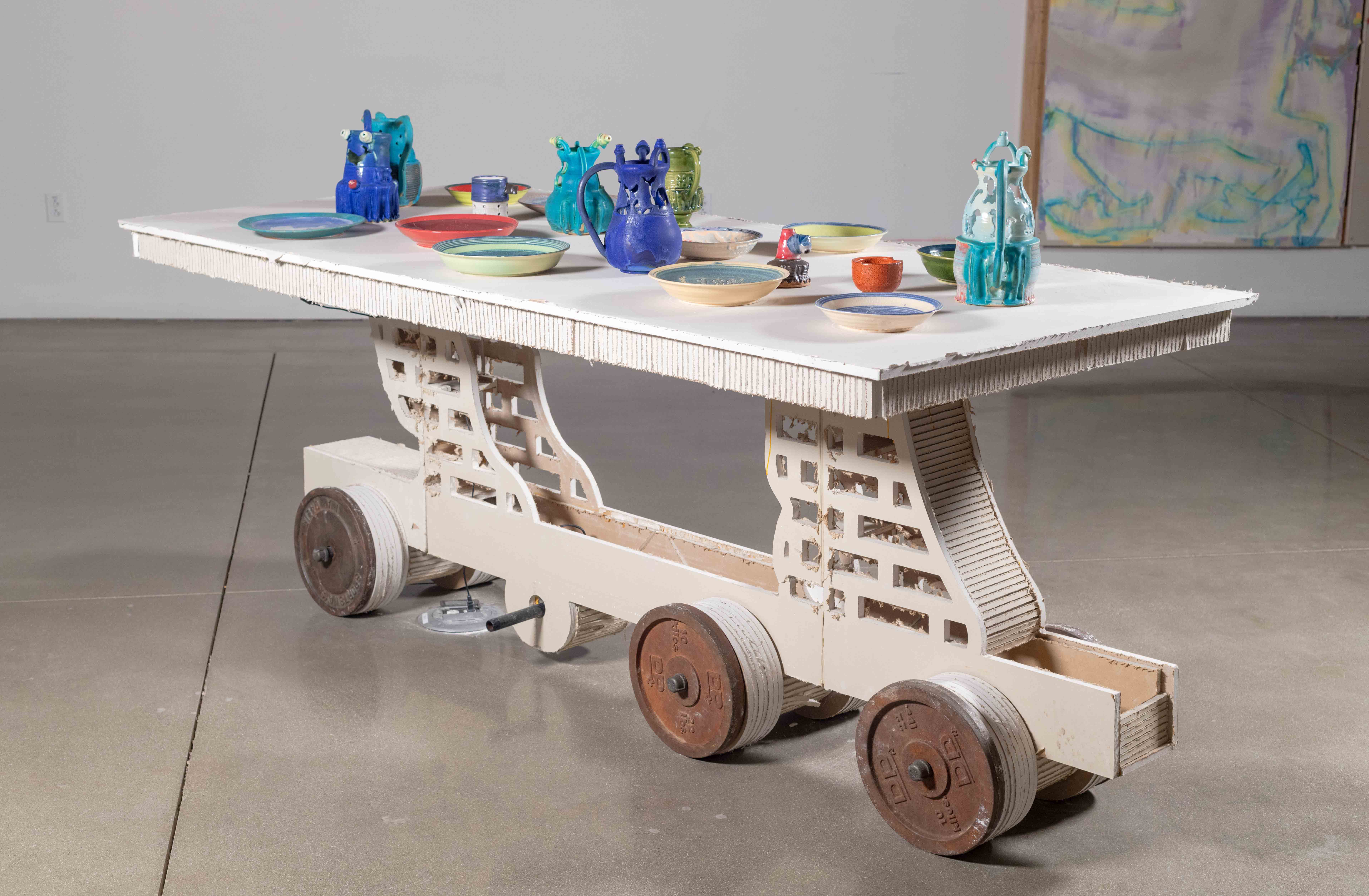 Drywall table with LED lights imbedded below the surface. 25 pound iron weights for
lifting are the tables wheels. The table legs are an outline of a horses feet. On top of the table
are an assorted gathering of brightly glazed Ceramic objects— Puzzle Jugs and WHO plates.