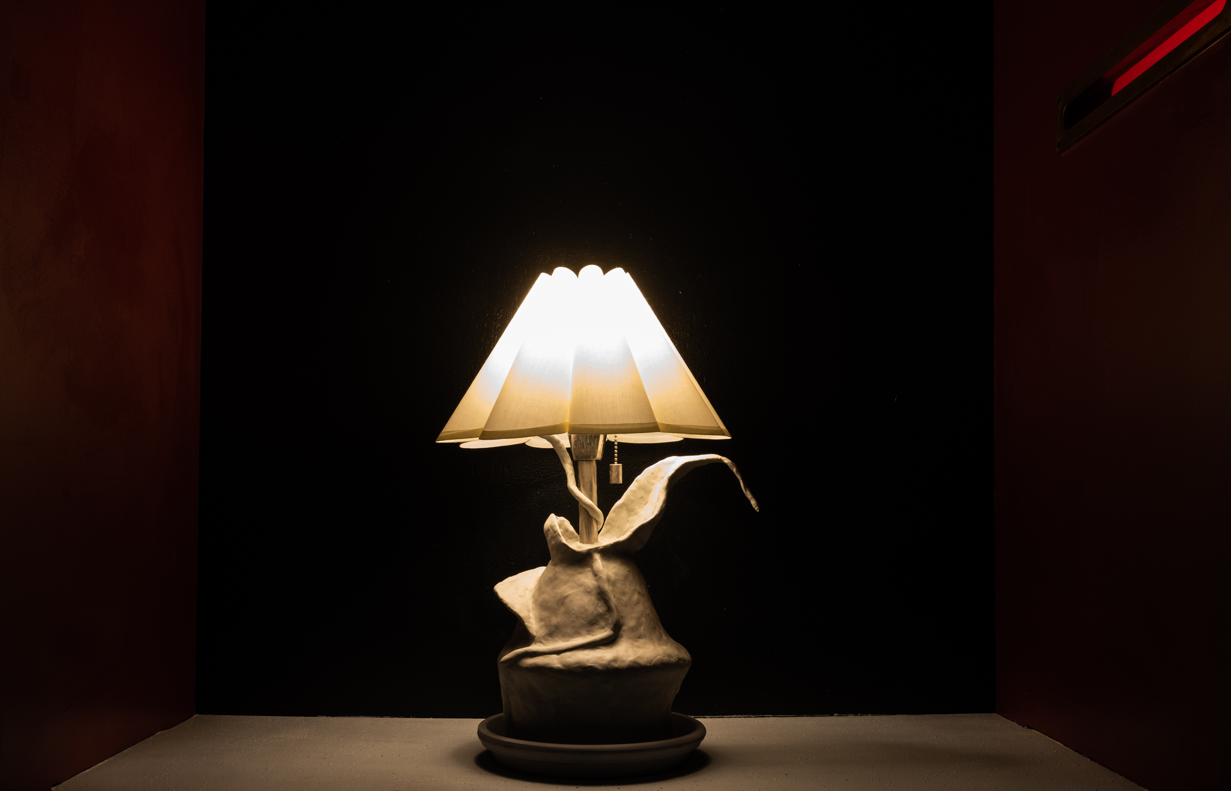This image is a detail shot of a hand-formed organically-shaped table lamp. The lamp is made from clay, paper maché, and epoxy resin. It is left untreated as a warm gray color. It is resting on top of a concrete pot draining dish. The lamp shade has folds reminiscent of petals. The lamp has “leaves” which grow out of the form. Off to the side is the mail slot with a red light glowing from within.