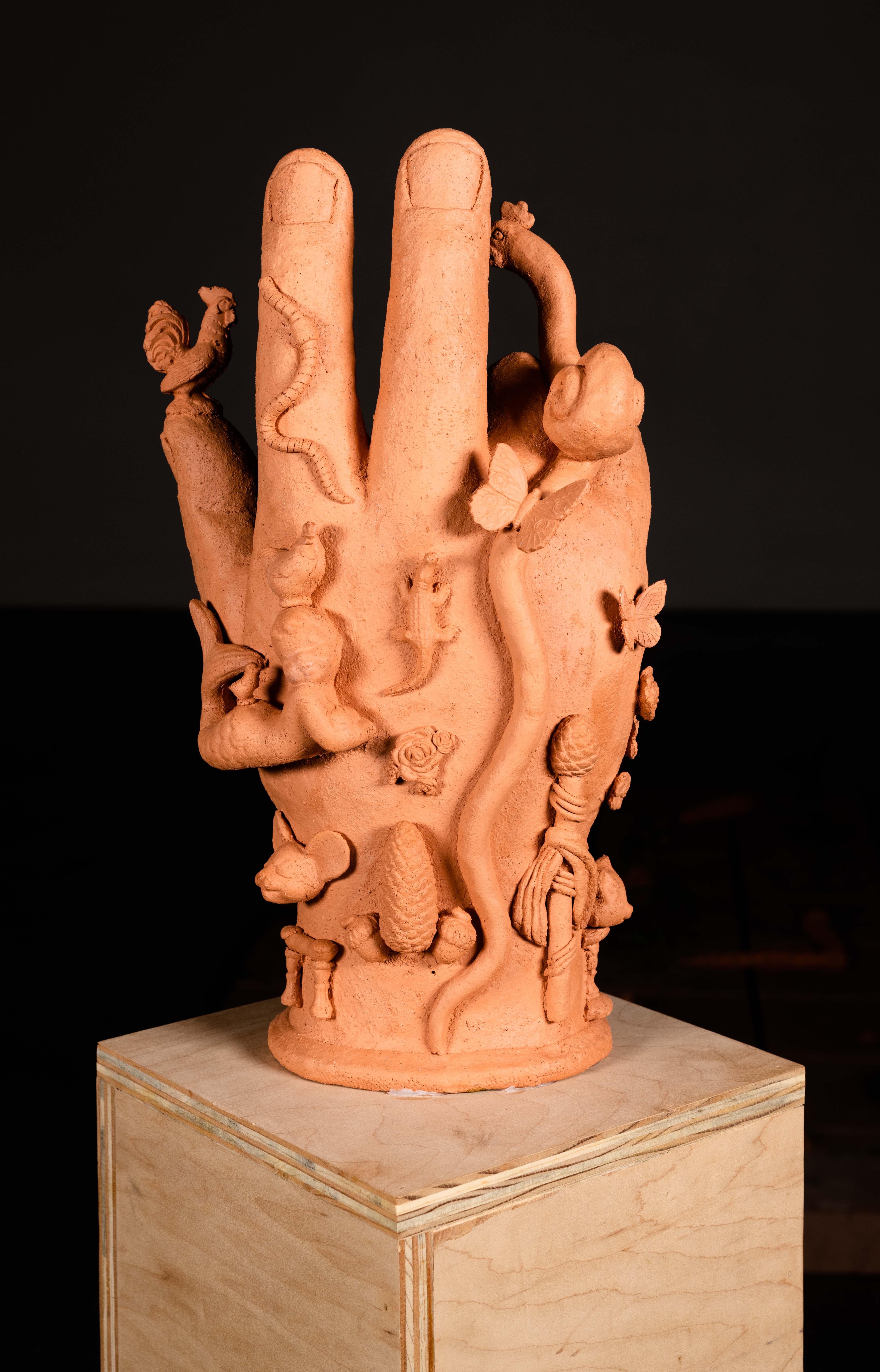 Detail of the back of a ceramic hand on top of a reclaimed wood pedestal. The hand  is in the position of the Hand of the Benediction. On top of the thumb is a small rooster. A  snail-like creature slides up the back of the hand and extends above the pinky and ring fingers.  A snail-shell and butterflies adorn its body. On the ride side of the hand is a phallic staff made  to look like an asparagus. In the center is a small lizard with roses below its tail as well as a  pine-cone with two acorns on either side. To the left of the lizard is a cherub-like figure with a  mermaids tail, a hen upon its head and a baby chick on its backside. Below the figure is a  mouse head above a small phallic arch structure. The museum wax holding the hand to the  pedestal is slightly visible.