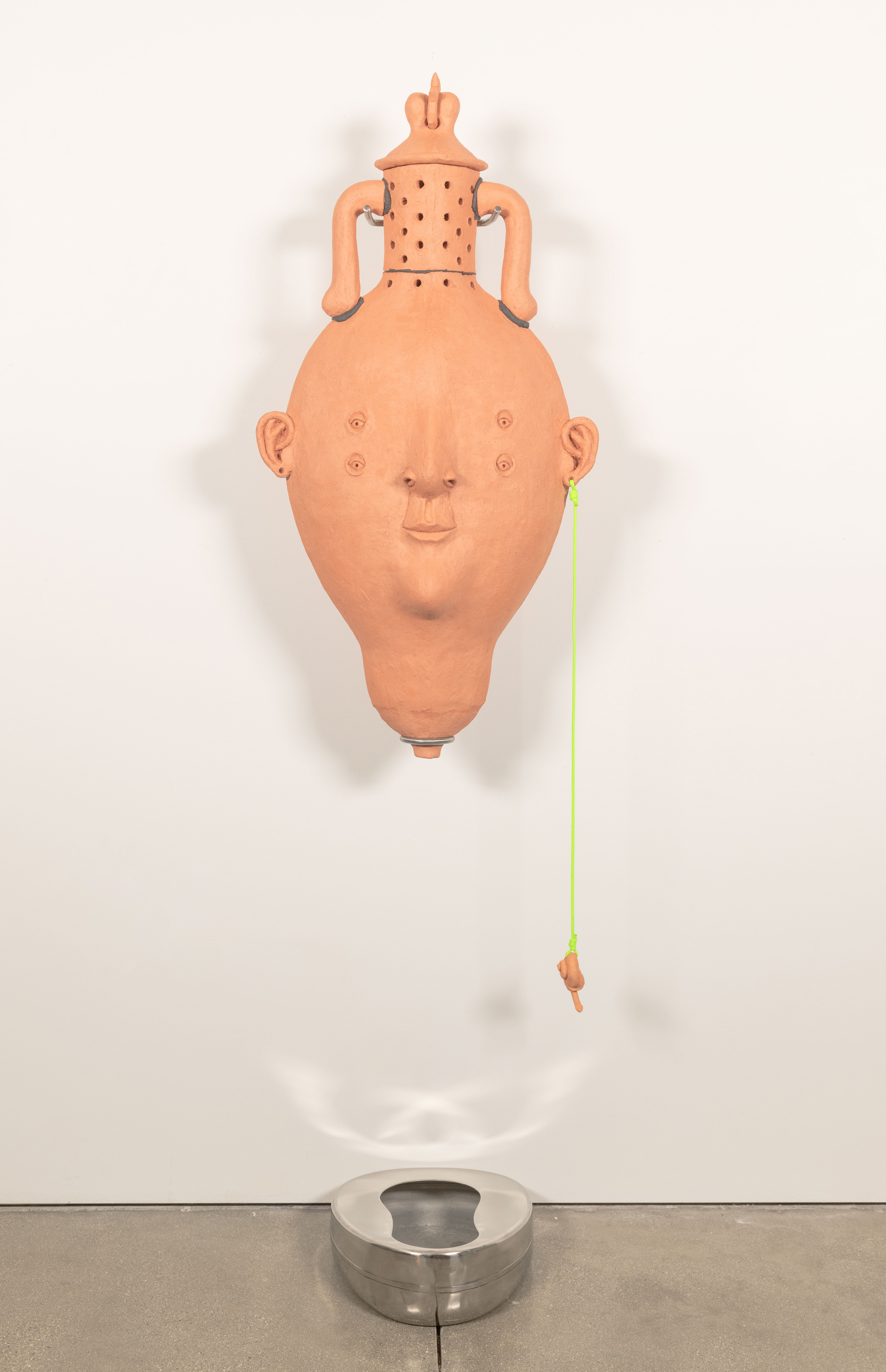 An anthropomorphic amphora hangs from the wall on a stainless steel mount
positioned over a bedpan. There is a lid and holes on the neck of the vase. Its base comes to a
point with a hole for drainage. From its ear hangs a snail-like charm on neon green Paracord.
Inside, red worms eat organic waste collected from my garden and the UCLA graduate studios.
Through the vermicomposting process a leachate is produced that collects in the bedpan
bellow.