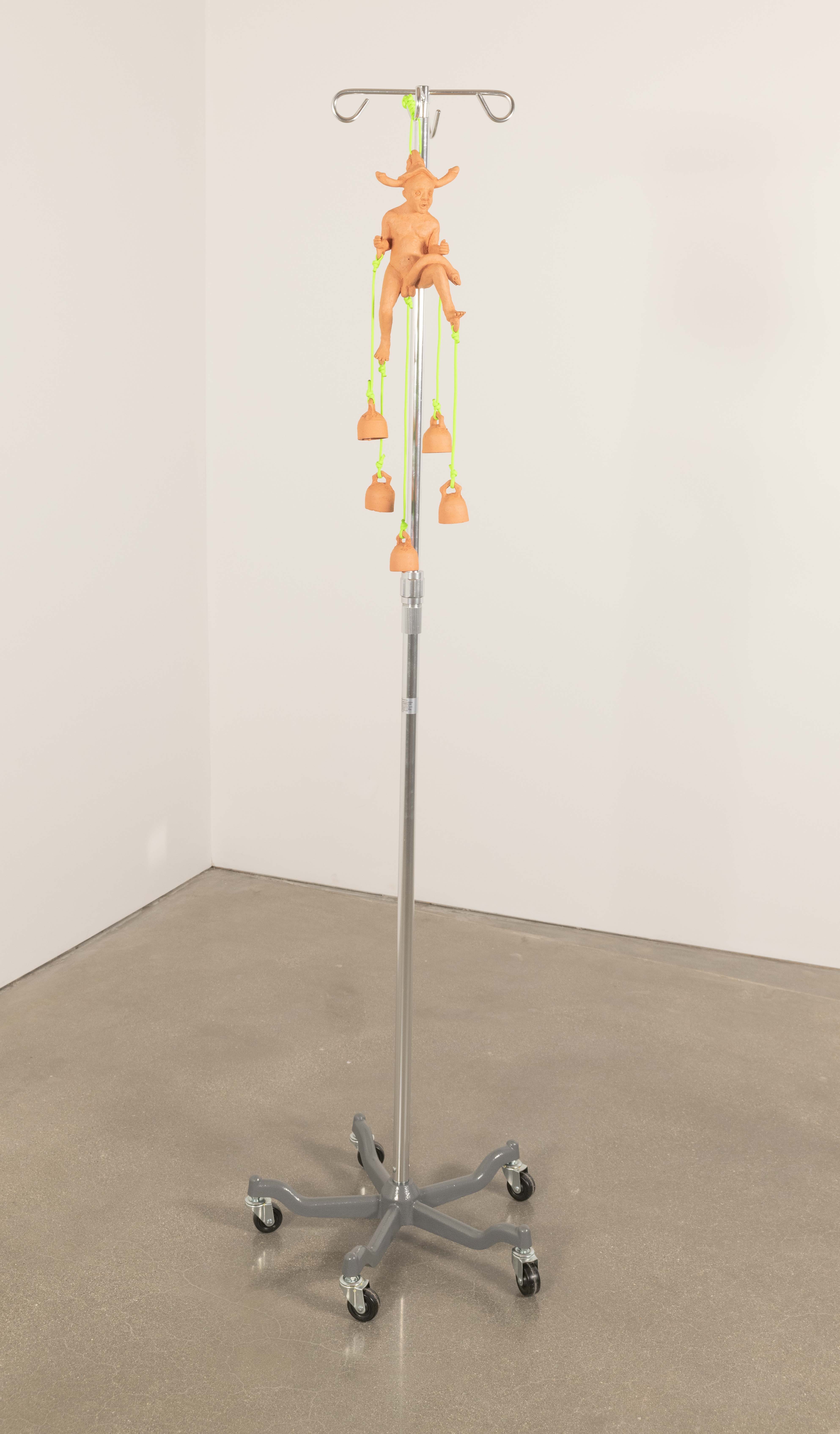 A ceramic wind chime composed of a nude figure with phallic bells attached to its
appendages with neon green Paracord hangs from a stainless steel iv pole.