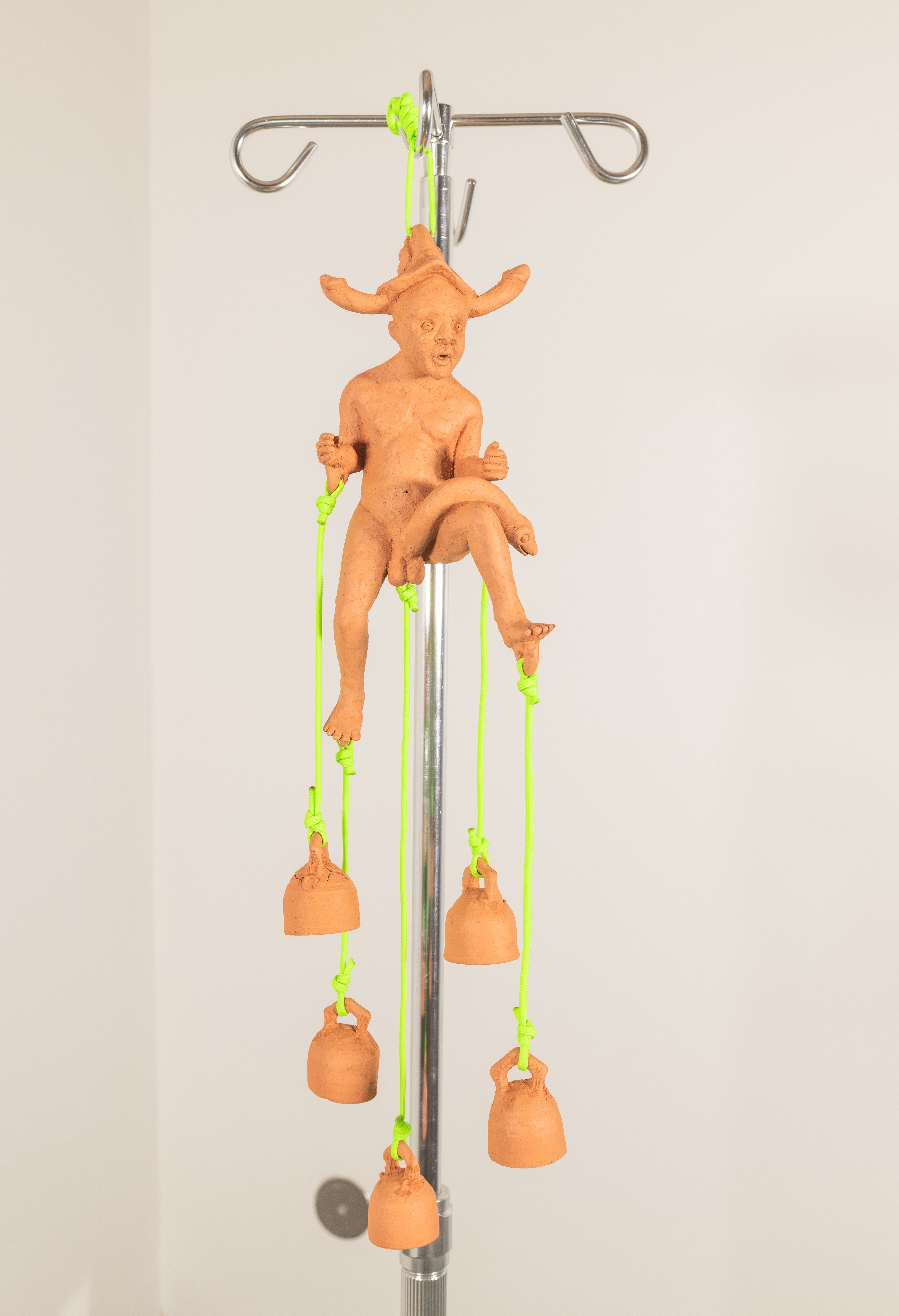 A ceramic wind chime composed of a nude figure with phallic bells attached to its
appendages with neon green Paracord hangs from a stainless steel iv pole.