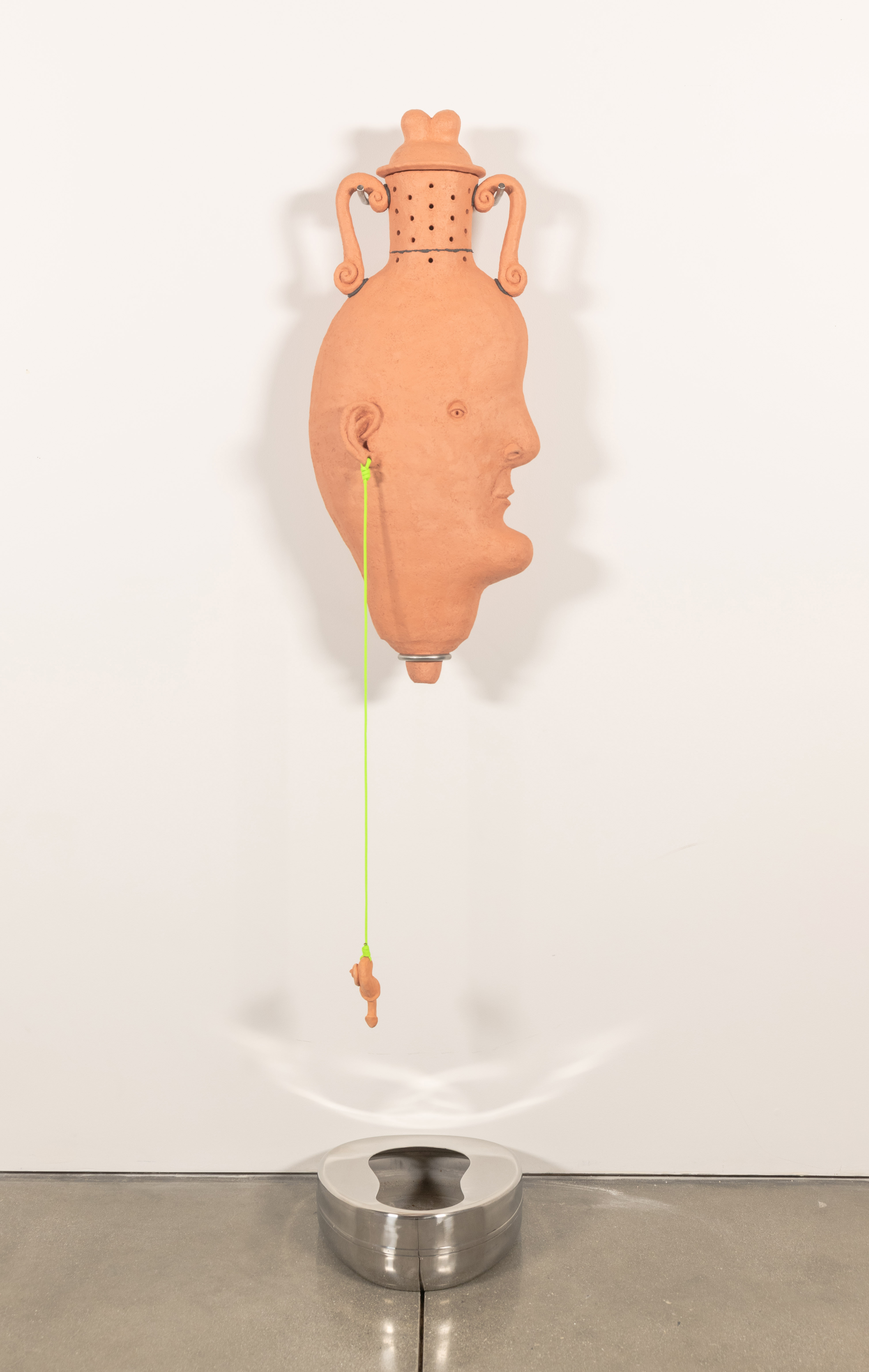 An anthropomorphic amphora hangs from the wall on a stainless steel mount
positioned over a bedpan. There is a lid and holes on the neck of the vase. Its base comes to a
point with a hole for drainage. From its ear hangs a snail-like charm on neon green Paracord.
Inside, red worms eat organic waste collected from my garden and the UCLA graduate studios.
Through the vermicomposting process a leachate is produced that collects in the bedpan
bellow.