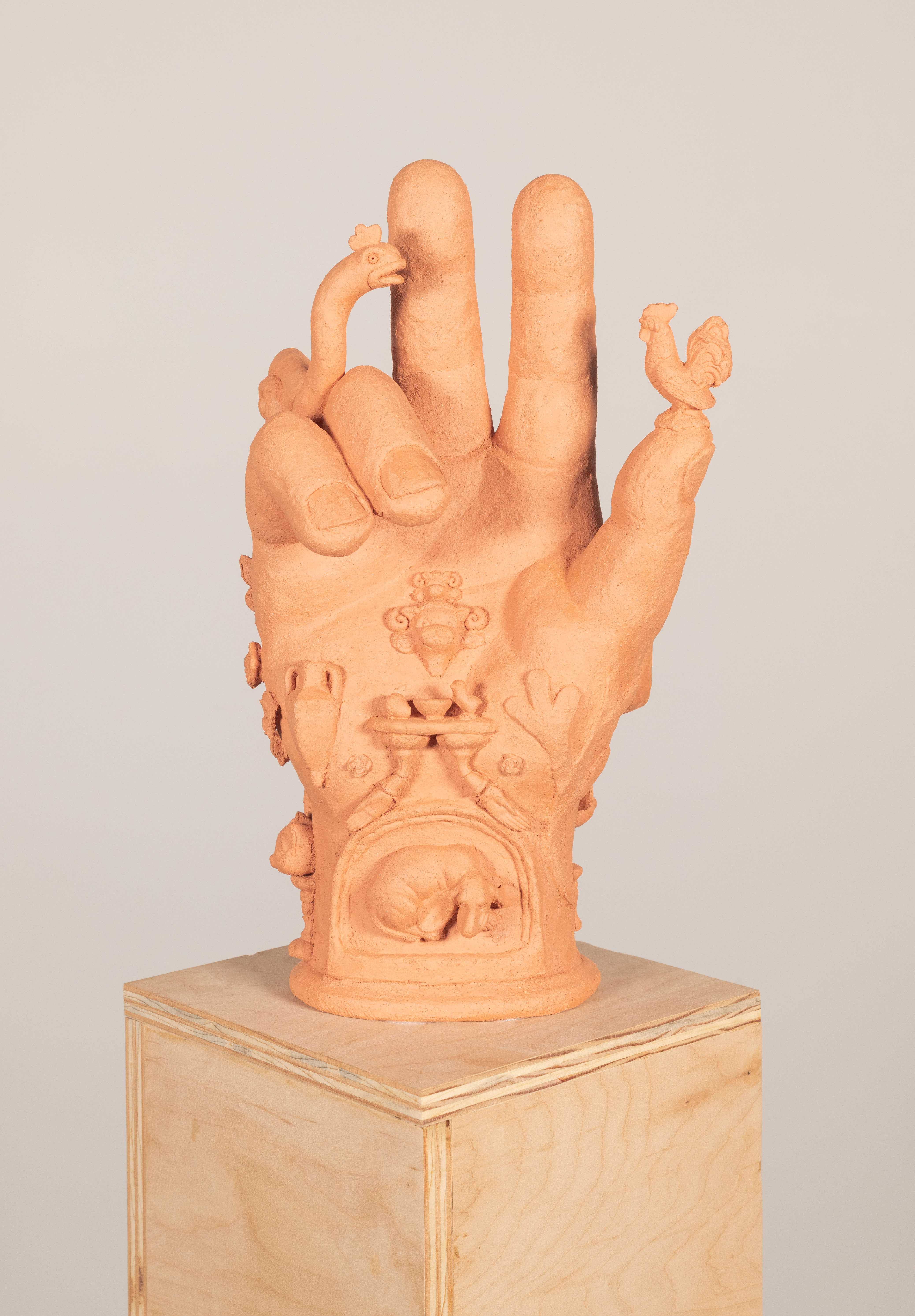 A ceramic hand on top of a reclaimed wood pedestal. The hand is in the position of
the Hand of the Benediction. On top of the the thumb is a small rooster. In the center of the
palm there are two goat-like heads. Below that is an offering table with a baby chick, bowl, and
acorn on its surface. The legs of the offering table turn into small outstretched hands that
frame a dog laying within a hearth on the wrist. There are floral and vegetal adoring the hand as
a well as a classical vase. A snail-like creature extends its neck from behind the pinky and ring
fingers. On the side of the wrist is a cats head resting upon a pediment. The museum wax
holding the hand to the pedestal is slightly visible. 