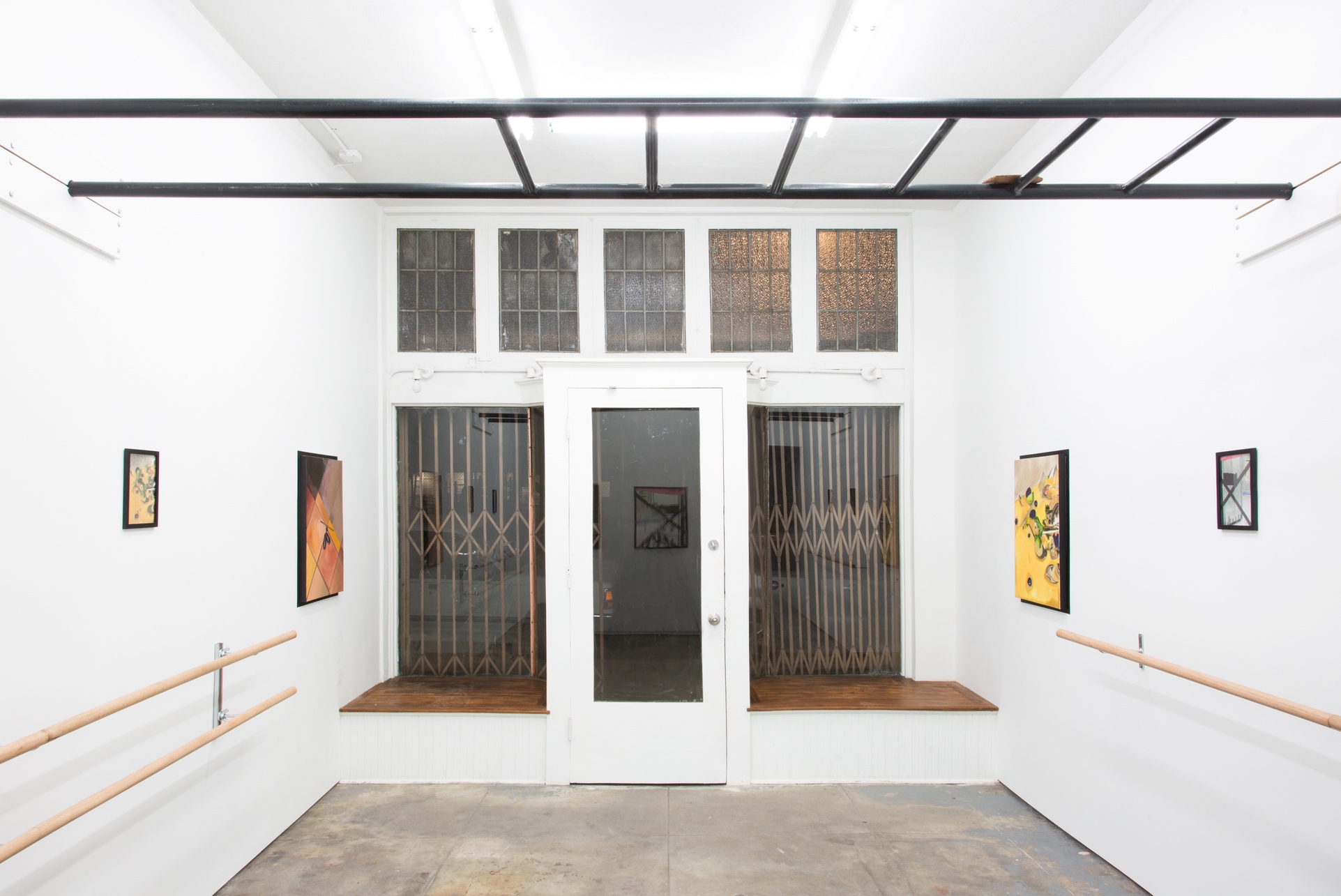 Installation image of Sparkling or still, an exhibition at In Lieu Gallery in Los Angeles. The picture is shot at a wide angle. The room is bright white with a gray concrete floor. There are two sets of ballet barres running along the walls and hung above them are 4 printed and framed images. The images are the reference photos for a series of four oil paintings that translate from the originals, also present within the room. Out-of-hand’s reach is a 300lb black steel monkey bar-like structure that runs from wall to wall. Resting on top of it is an unfired clay tablet which sits precariously on top of the structure.  The clay tablet has the 10 Commandments scratched onto the front and the back of the surface.