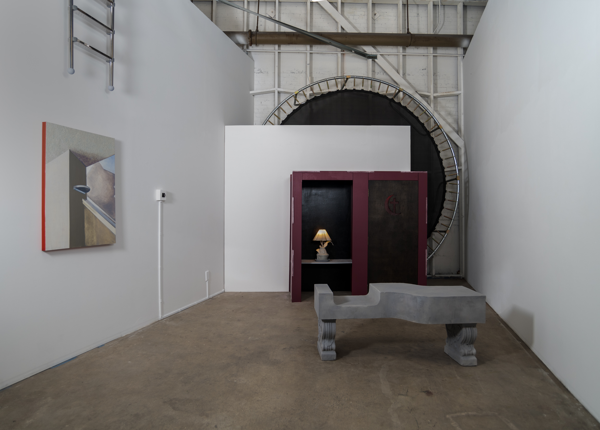  This is an Installation image of I/meld/a/Spirit, the culminating thesis work by Cherisse Gray. I/meld/a/Spirit is a sculptural installation situated in a trapezoidal room with no ceiling. Against the far wall is a 15 foot trampoline oriented vertically and sandwiched between the existing wall of the building and a wall the artist built. On the other end of this “sandwich” is a dark maroon confessional booth. On the left hand side (for the confessor) is a lit gray lamp shaped as an organic-looking form. In front of the booth is an undulating concrete bench with an ergonomic dip. To the left of the bench is an audio knob that controls a three minute sound piece which loops on the inside of the priest's side of the booth. The knob can be turned by the viewer to effectively "turn on" or "turn off" the voice of the artist. A crimson red light emanates from the priest’s side of the booth. To the left of the knob is a painting and above the painting is an aluminum pool ladder that leads only to the other side of the wall. The ladder is out of reach for all viewers. The booth cannot be entered.