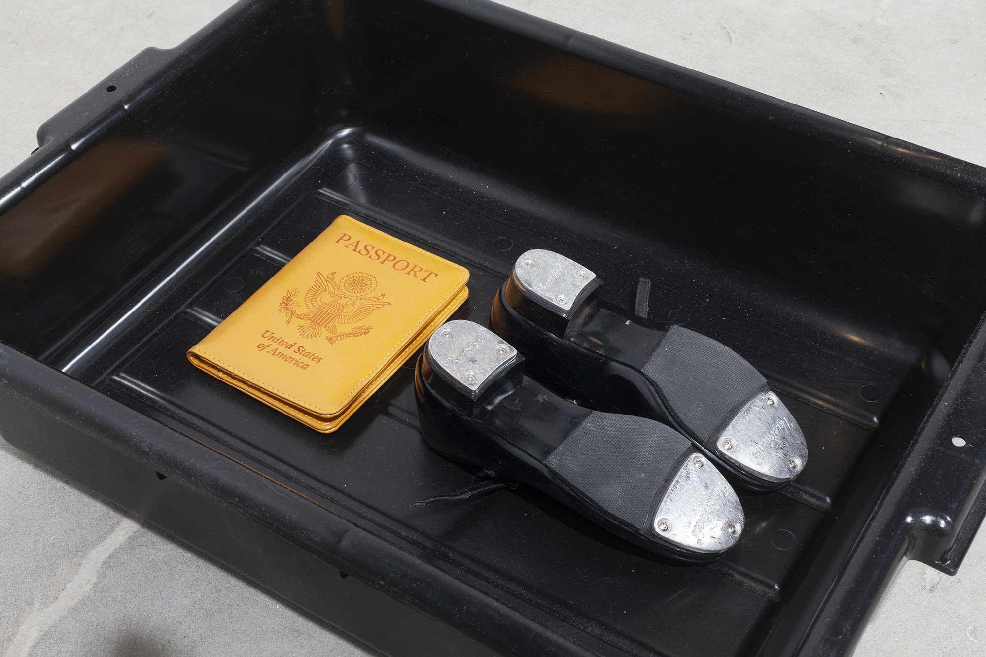 Detail image of a black TSA bin sitting on the floor. Inside of it are the following items: a yellow American passport holder and a pair of children’s tap shoes.
