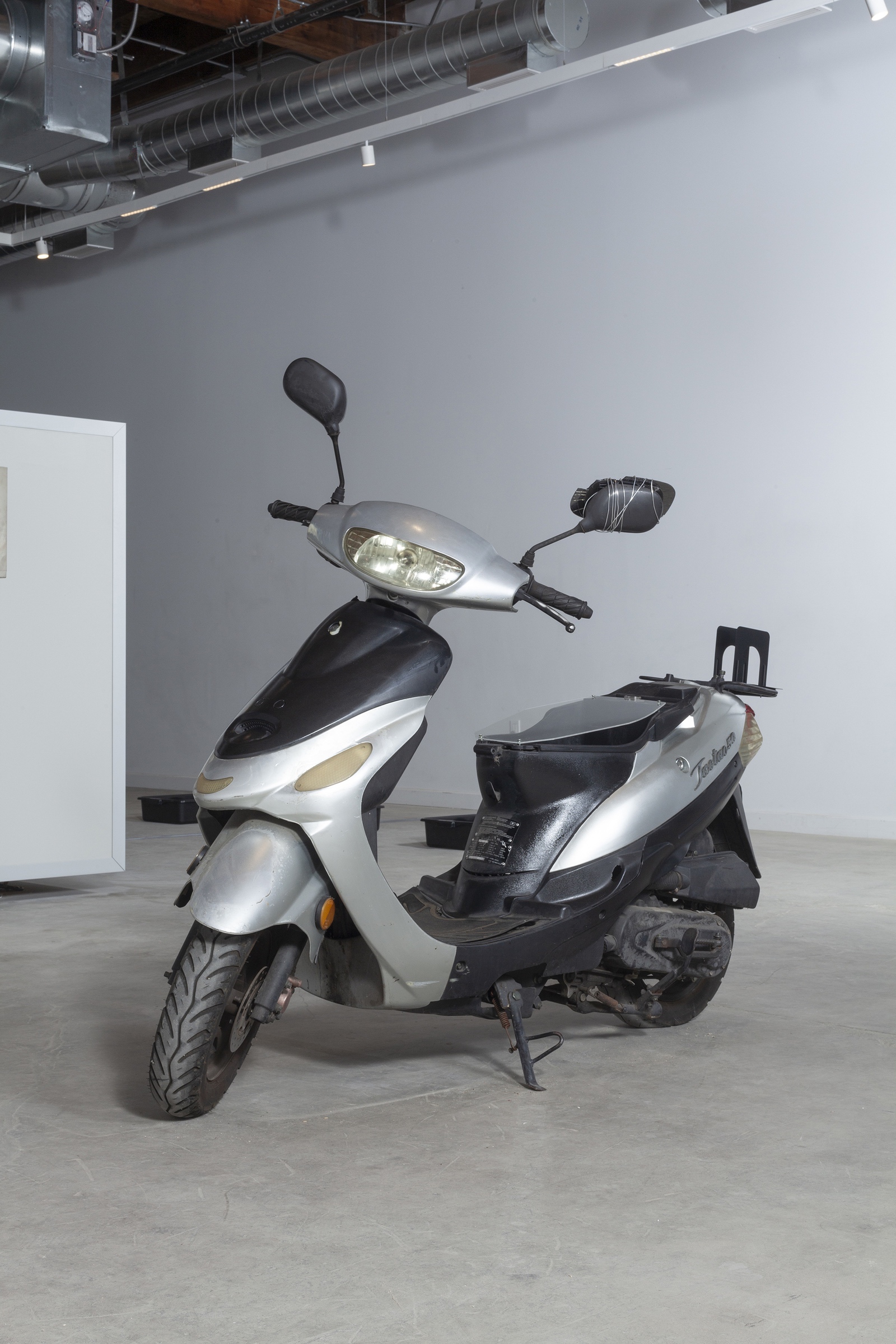 Installation image of  a parked scooter. The storage compartment contains a stack of thick documents made inaccessible through a sealed plexiglass barrier.