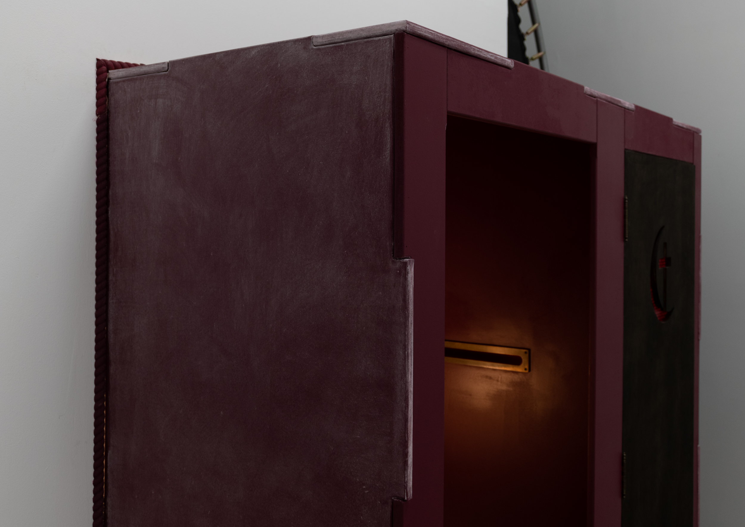This image is a detail shot of the side of the confessional booth. The edges of the maroon confessional booth are treated with a light patina of white chalk, separating it from the front which is still "clean". The warm glow of the table lamp is reflected onto the interior wall. The booth is held together through a series of finger joints.