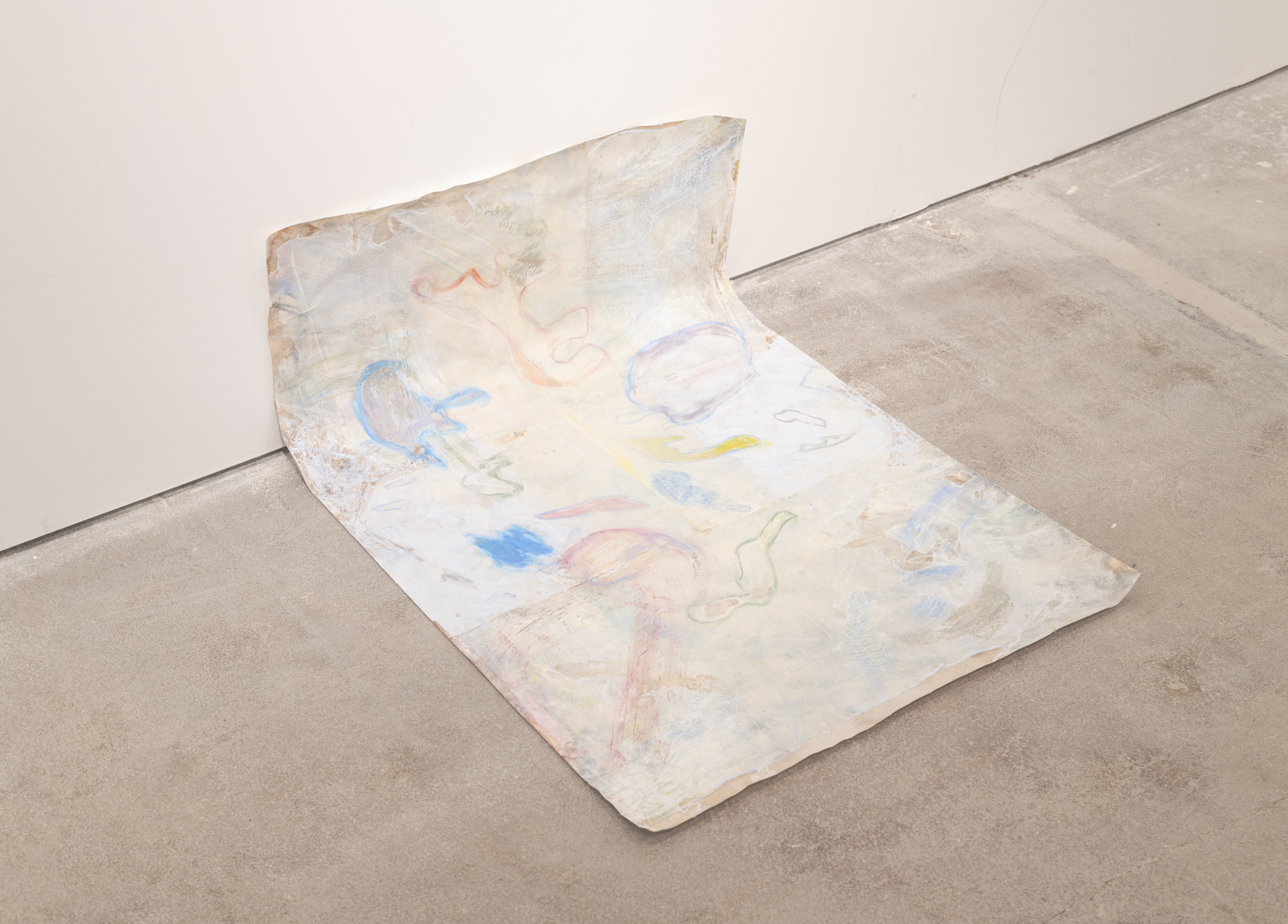 Paper sits against the wall with the top third of it sitting against the wall while the rest sits on the floor. Background possesses washed out walnut ink with a layer of cream colored acrylic paint. Residual watermarks lingering are outlined and filled in with various pastel colors--chartreuse, various blues, burgundy, forest green, and red-orange. The overall shapes are obscured by a foggy layer of white that's slightly translucent.
