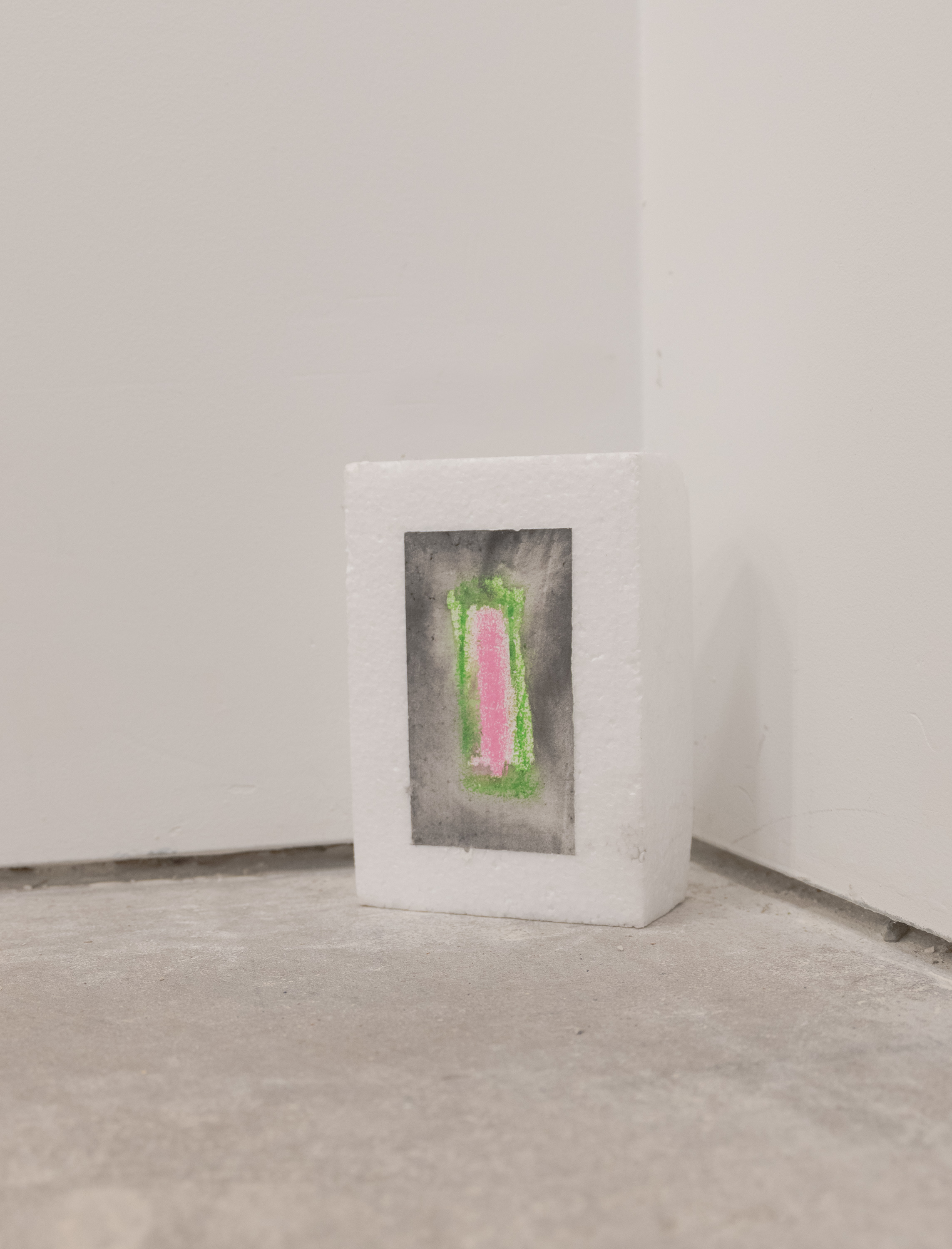 A block of styrofoam rests in the corner of the room touching the floor. It consists of a pink center with a green outline. Outside the green is a gradient of color from gray to black. The darkest part sits closest to the edge.