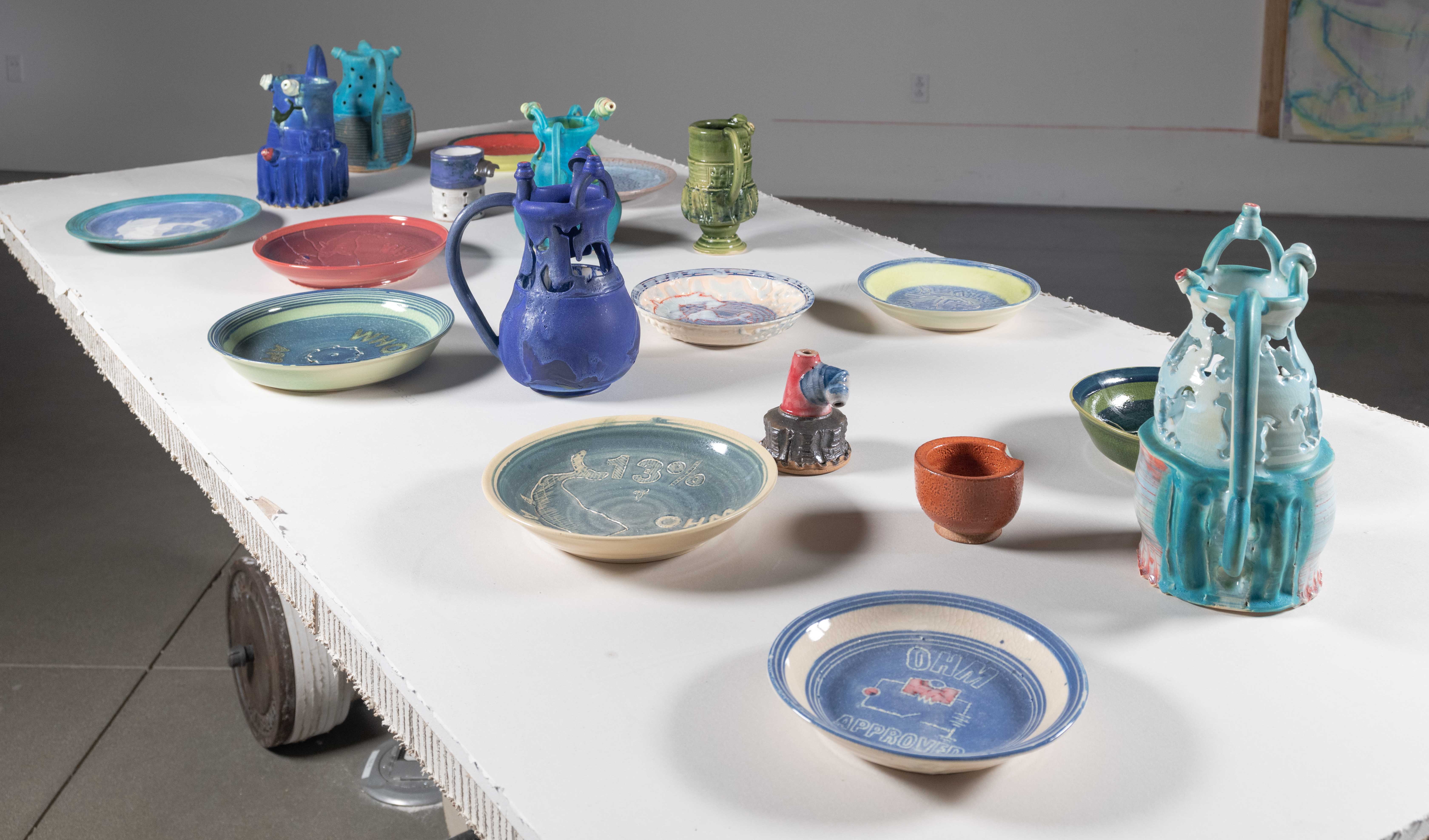 Drywall table with LED lights imbedded below the surface. 25 pound iron weights for
lifting are the tables wheels. The table legs are an outline of a horses feet. On top of the table
are an assorted gathering of brightly glazed Ceramic objects— Puzzle Jugs and WHO plates.