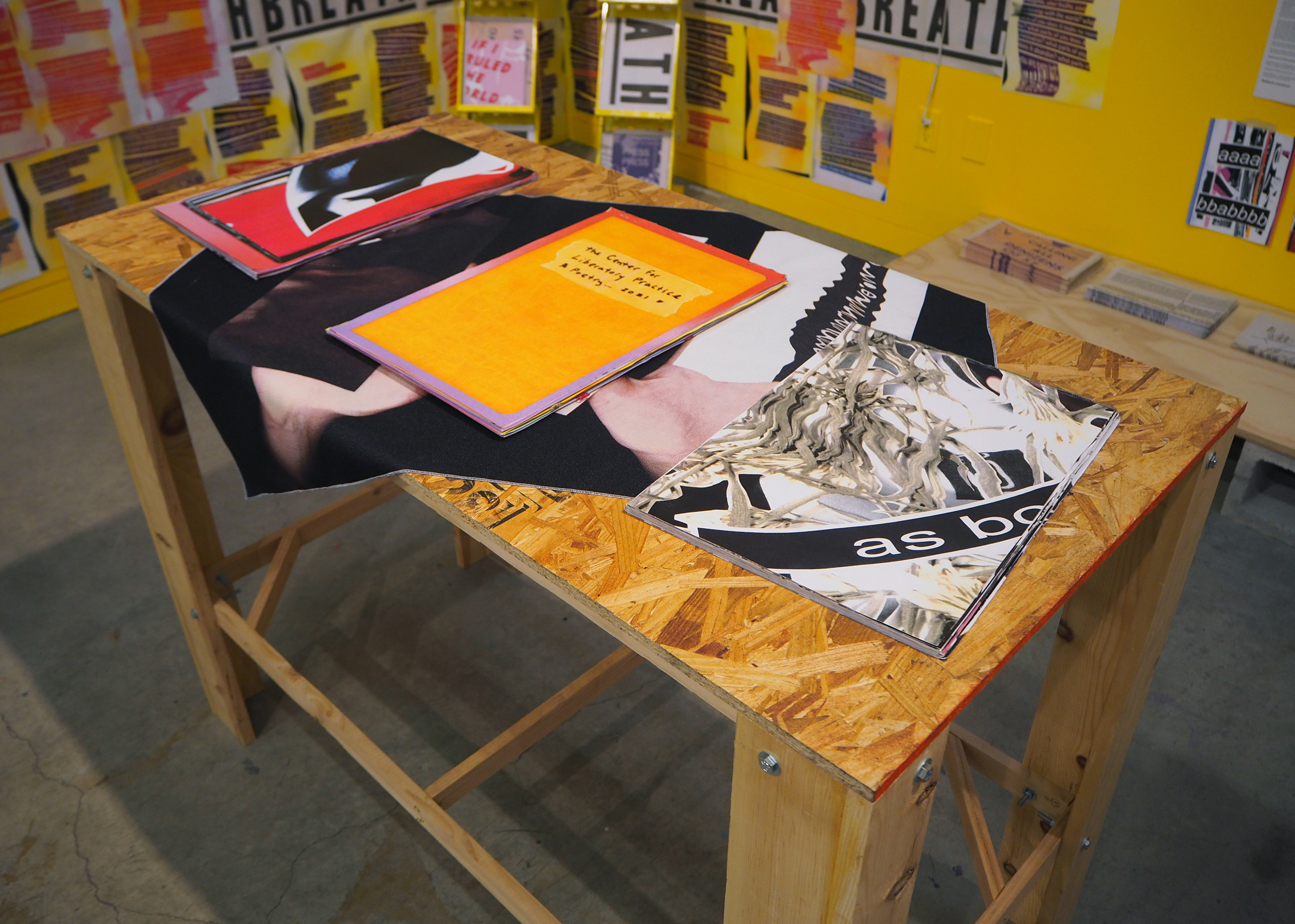 Three books sit on a table. The table top is made of OSB with a fabric draping over its center diagonally. The left and right sides of the tabletop are revealed. The fabric is white and black and feels like a xerox print or scan of warped lettering and hands. The book on the most left side is red and black, the middle book is yellow, and the book on the right has scans of light green plants and a black line with a few white letters overtop that say, “as bo” and are cut off. 