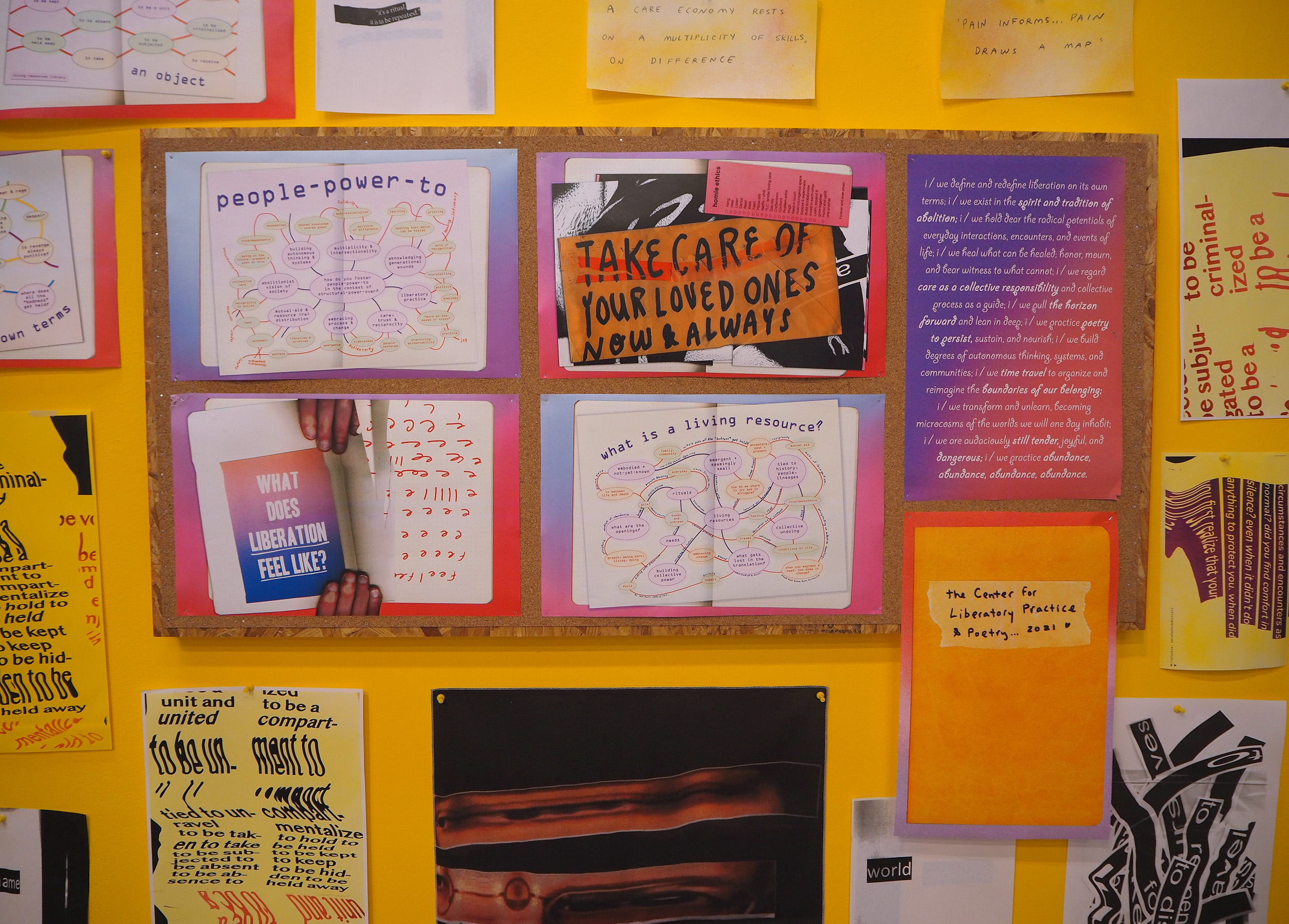 A corkboard covered in prints sits on top of a yellow wall. The yellow wall has prints hung up with yellow push pins. The cork board has four horizontal prints of open notebook pages and mind maps that brainstorm the Center for Liberatory Practice & Poetry. Most legible from this picture are two pieces: the first is two hands holding a card with a pink and blue gradient that reads, “what does liberation feel like?”. The second most legible image is of an envelope with handwritten text that reads, “take care of your loved ones now & always”. 
