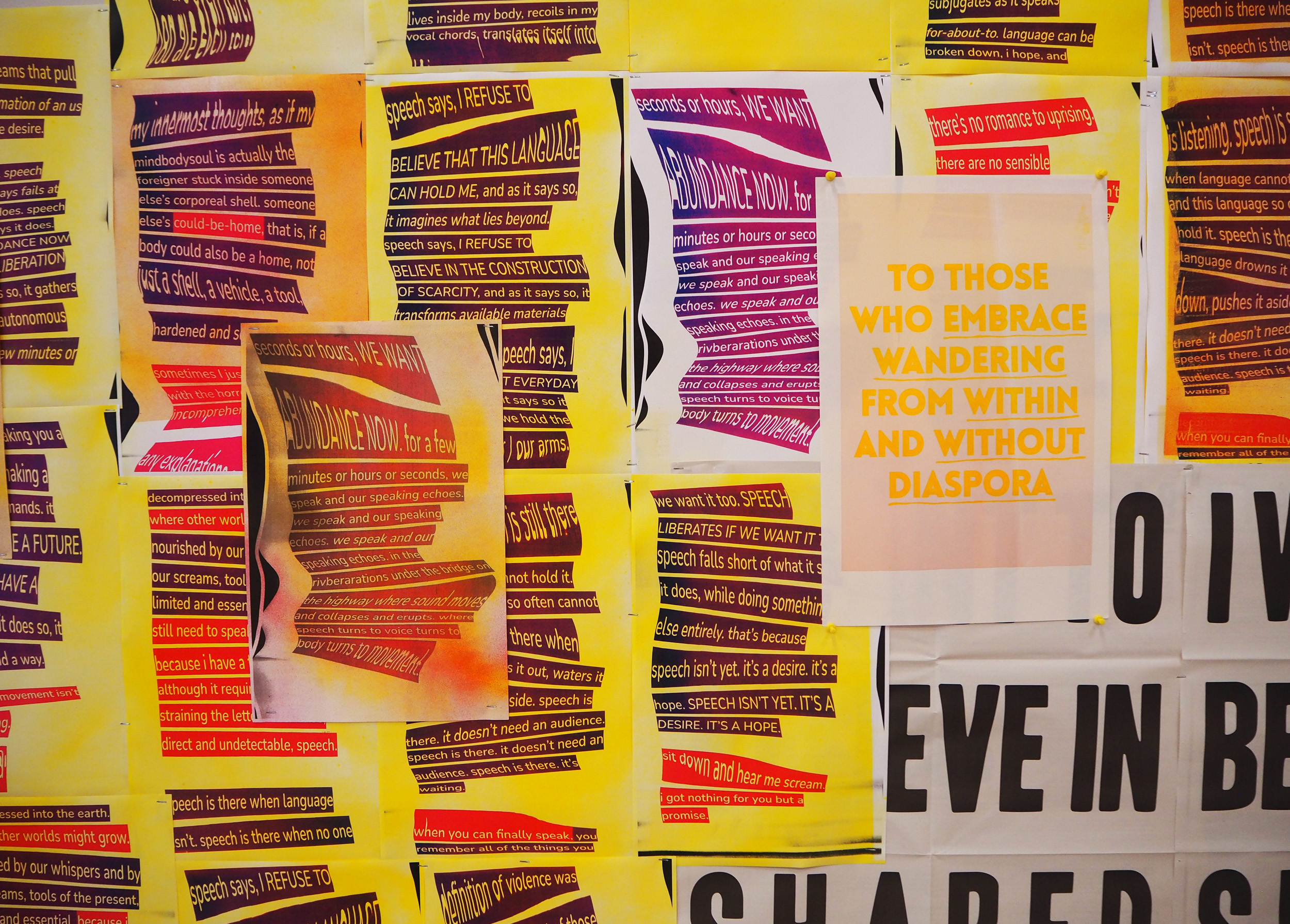  A close up of a wall of posters. The posters have yellow, orange, and white backgrounds with blocks of warped text overtop. The text blocks range in purple to blue shades with yellow and white letters. Most visible text reads, “abundance now”. The posters all look similar to one another excluding one poster with much clearer text that reads, “To those who embrace wandering from within and without diaspora” in bright yellow-orange bold letters, over a pink background. This poster is put up with pushpins rather than stapled like the rest. 
