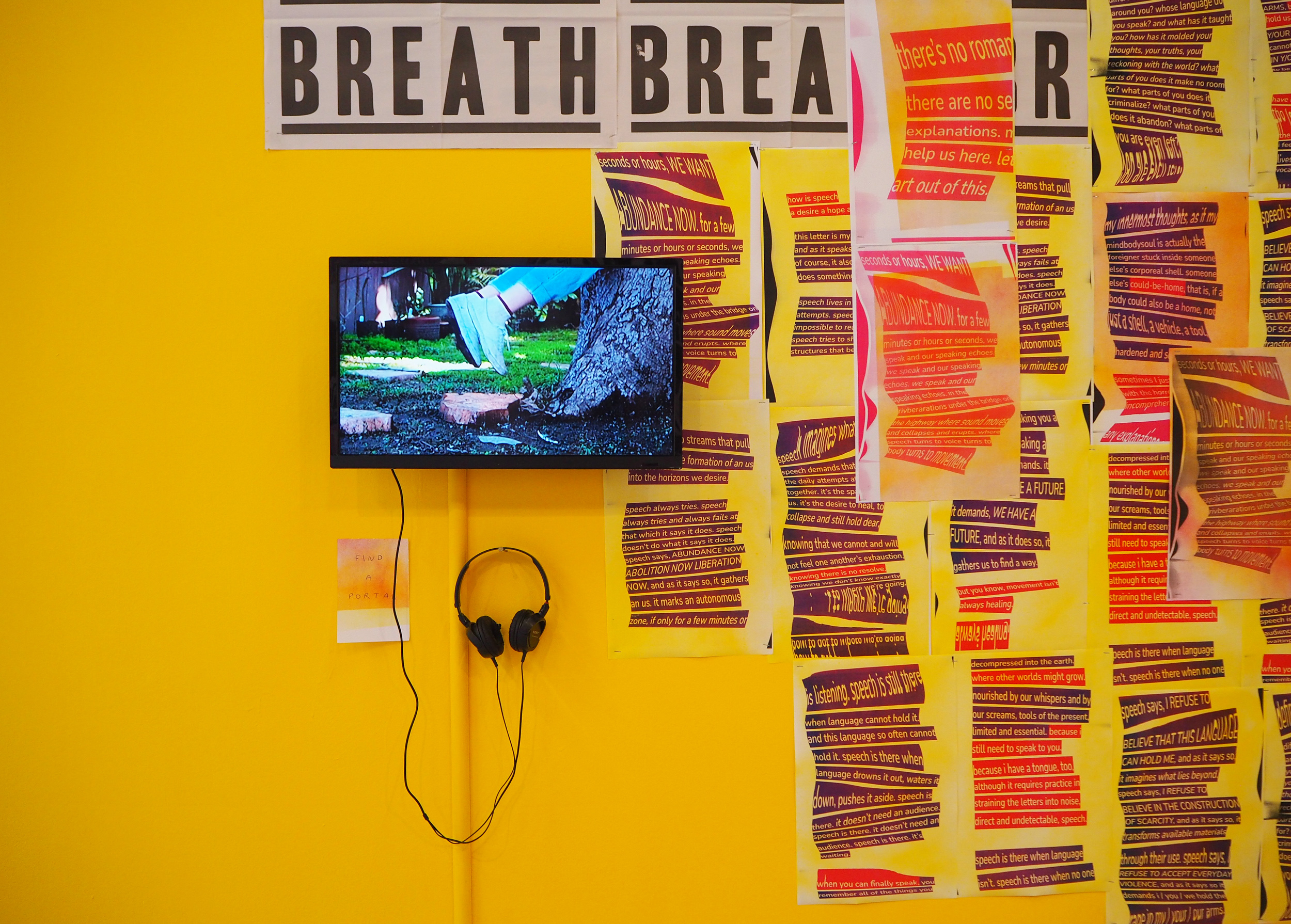 A yellow wall with a few posters on the right side. There is a screen hung on the middle of the wall with headphones that hang underneath. On the screen we see legs floating near the trunk of a tree. There are shades of green, blue and grey which sharply contrast with the yellows, pinks, and purples that surround. Above the screen we see the bottom of large posters that are hanging over top that read, “breath, breath”. 
