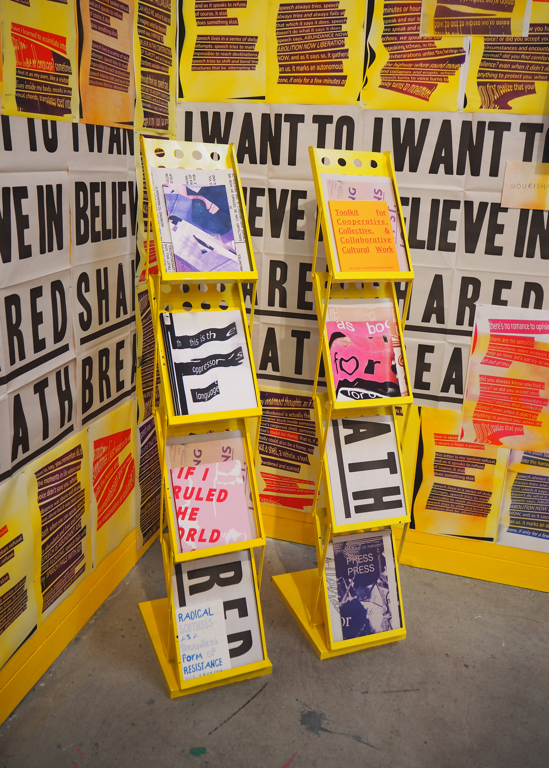 Two metallic, yellow publication stands sit at the corner of two walls. The walls are covered in grey and yellow posters. The publication stands are structured like accordions and have different books in each section. They have four sections to put books and magazines in. They are colorful and each section looks different. 
