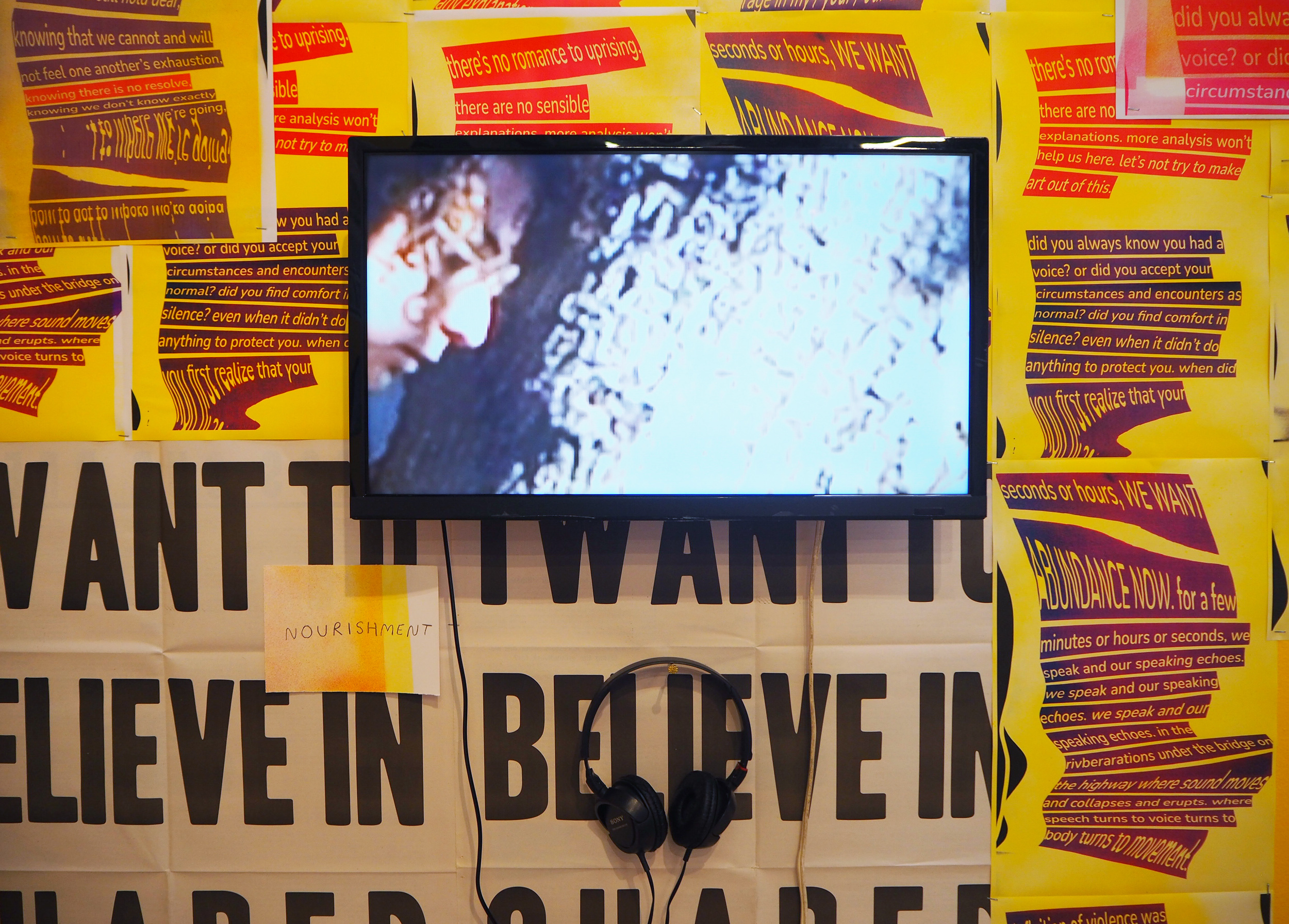 a screen hangs on a wall covered in various prints. The screen shows the image of a person’s face looking downward, very close to the trunk of a tree. The person is whispering to the tree. 