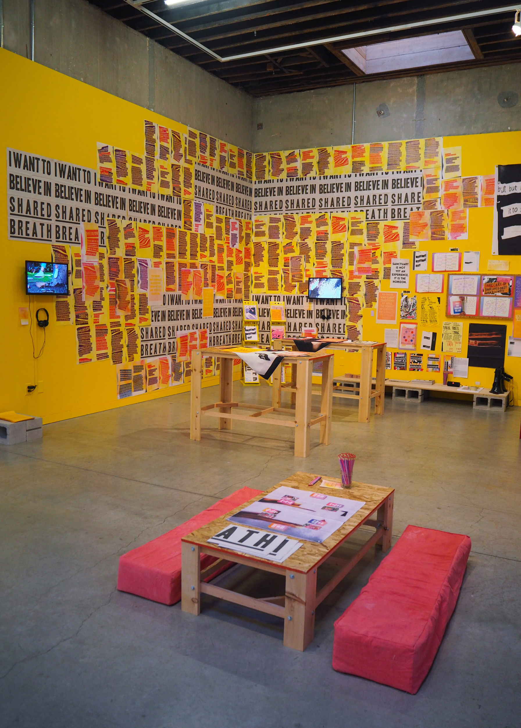 Two yellow walls covered in pink, purple, and grey prints surround a series of tables. One table is lower to the ground and sits in the foreground of the image. It is surrounded by two long, pink floor cushions. There are two small screens on the walls, one on the left and one on the middle right. The space feels busy. The floor is grey and slightly reflects the yellow of the walls. 
