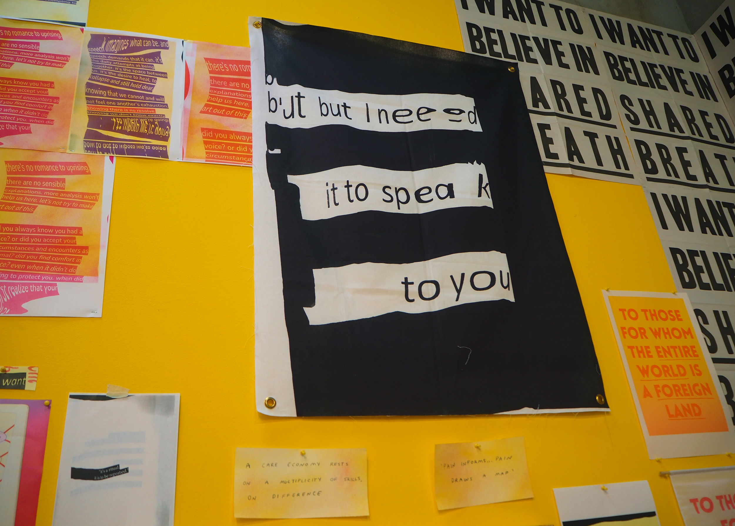 A close of one of the pieces on the yellow wall. The image shows a black and white banner with golden grommets hanging. In warped and somewhat legible text, the banner reads, “but i need it to speak to you” in black letters over top blocks of white, which sit over top a mostly black background. 
