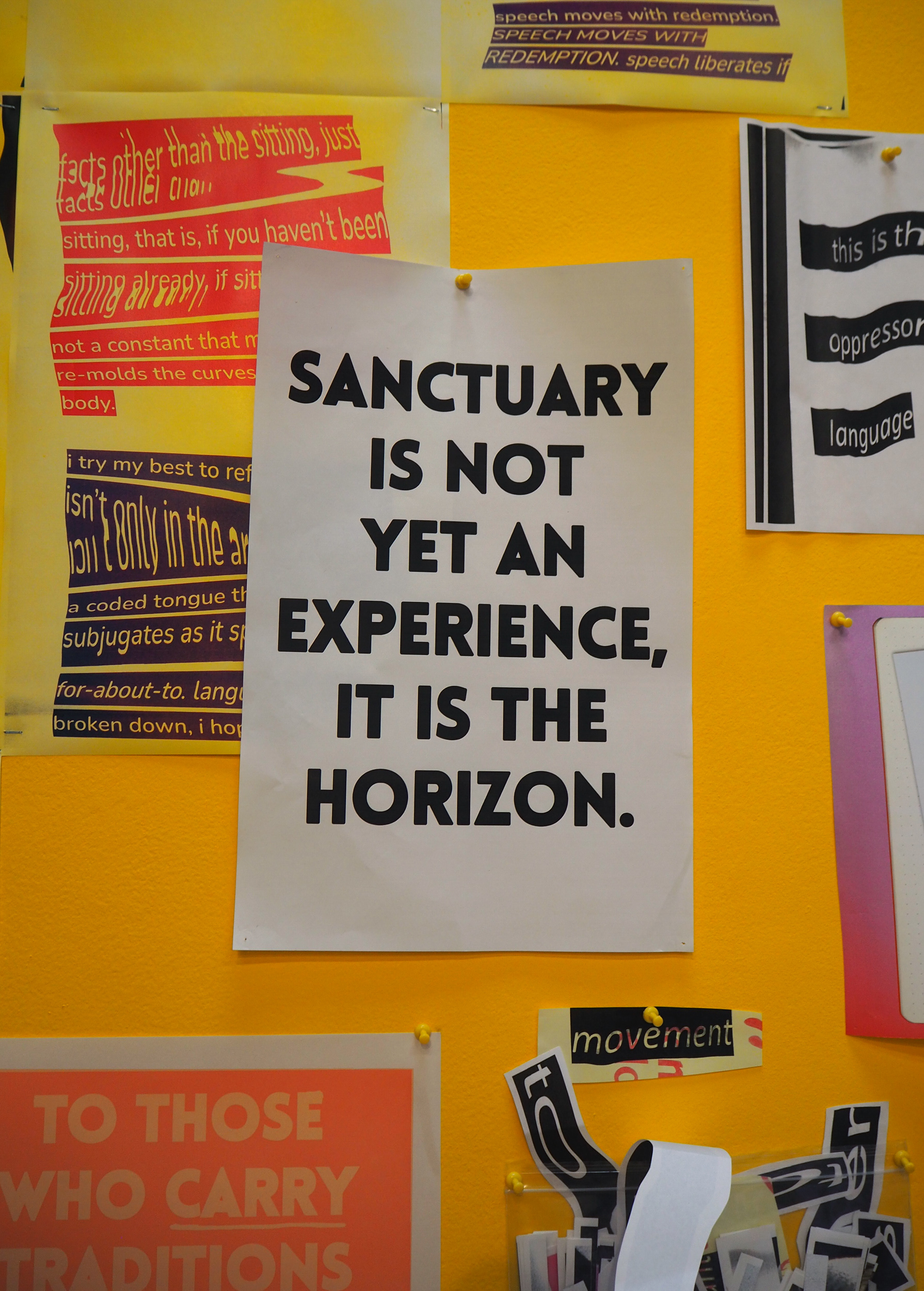 A white print is hung on a yellow wall with a yellow pushpin. In bold letters it says, “Sanctuary is not yet an experience, it is the horizon.” Underneath it is a small cut out that says, “movement” also put on the wall with a yellow pushpin. 
