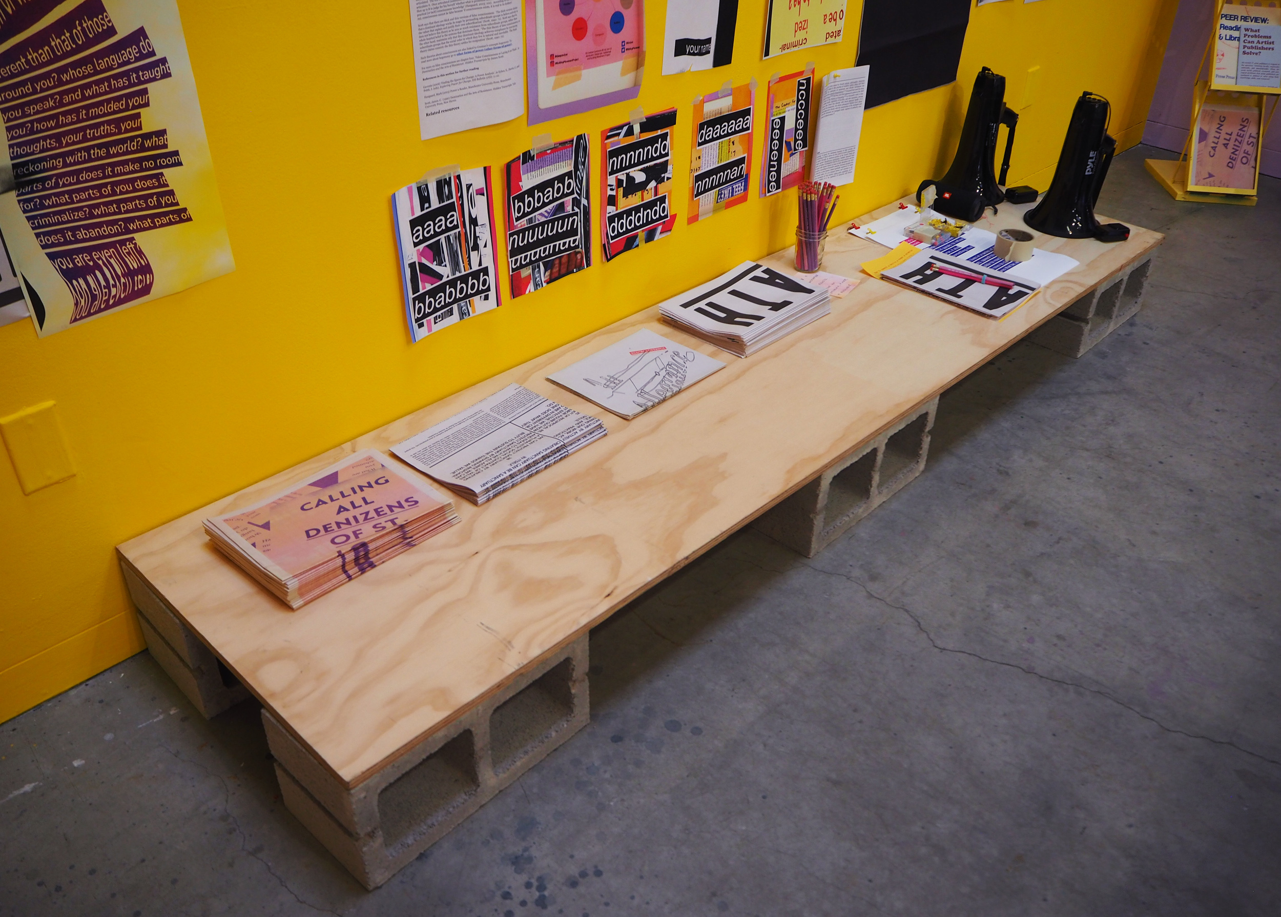A wooden shelf sits on the floor on top of concrete blocks. It has a few stacks of newspapers on top of it and two black megaphones sitton the edge. The wall behind it is yellow. 
