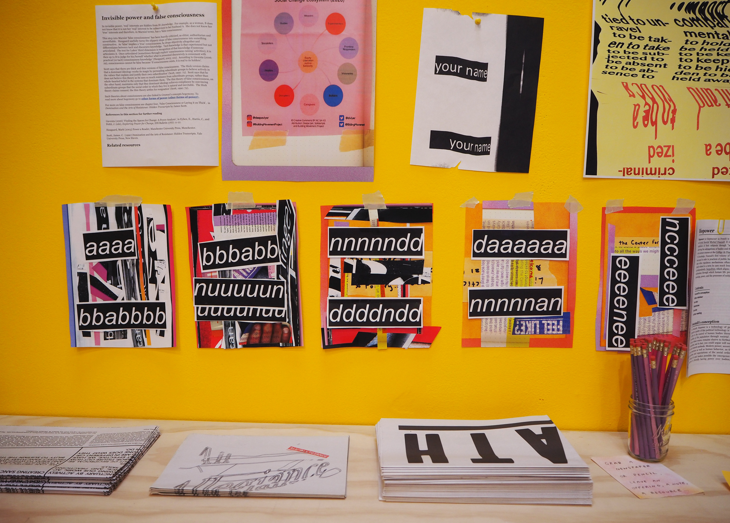 Five colorful collages are taped to a yellow wall. The same shelf sits below with piles of newspapers and a jar with pink and purple pencils. The collages have shades of purple, pink, white and black. They have a few strips of black with white letters repeated on top. Together, the pieces read, “aaaaaabbaabbbbuuuunnnndddddaaannnccceeeecccceeeee”. 