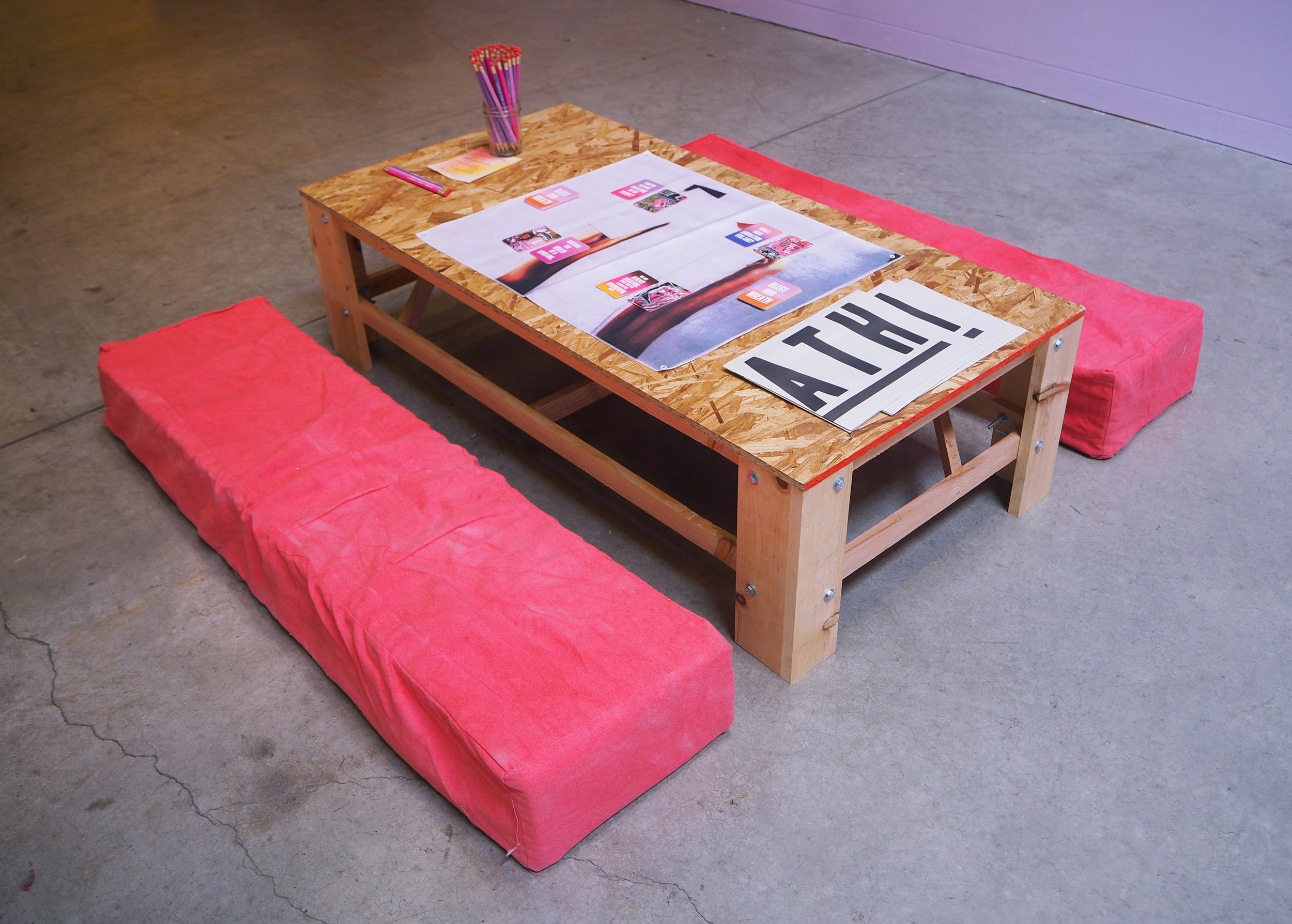  A wooden table sits lower to the ground. It is surrounded by two long, pinkish floor cushions. The table looks like a work table. It has cards laid out on it on to top of a whtie and grey fabric. The floor is grey and we see a glimpse of a purple wall behind. 
