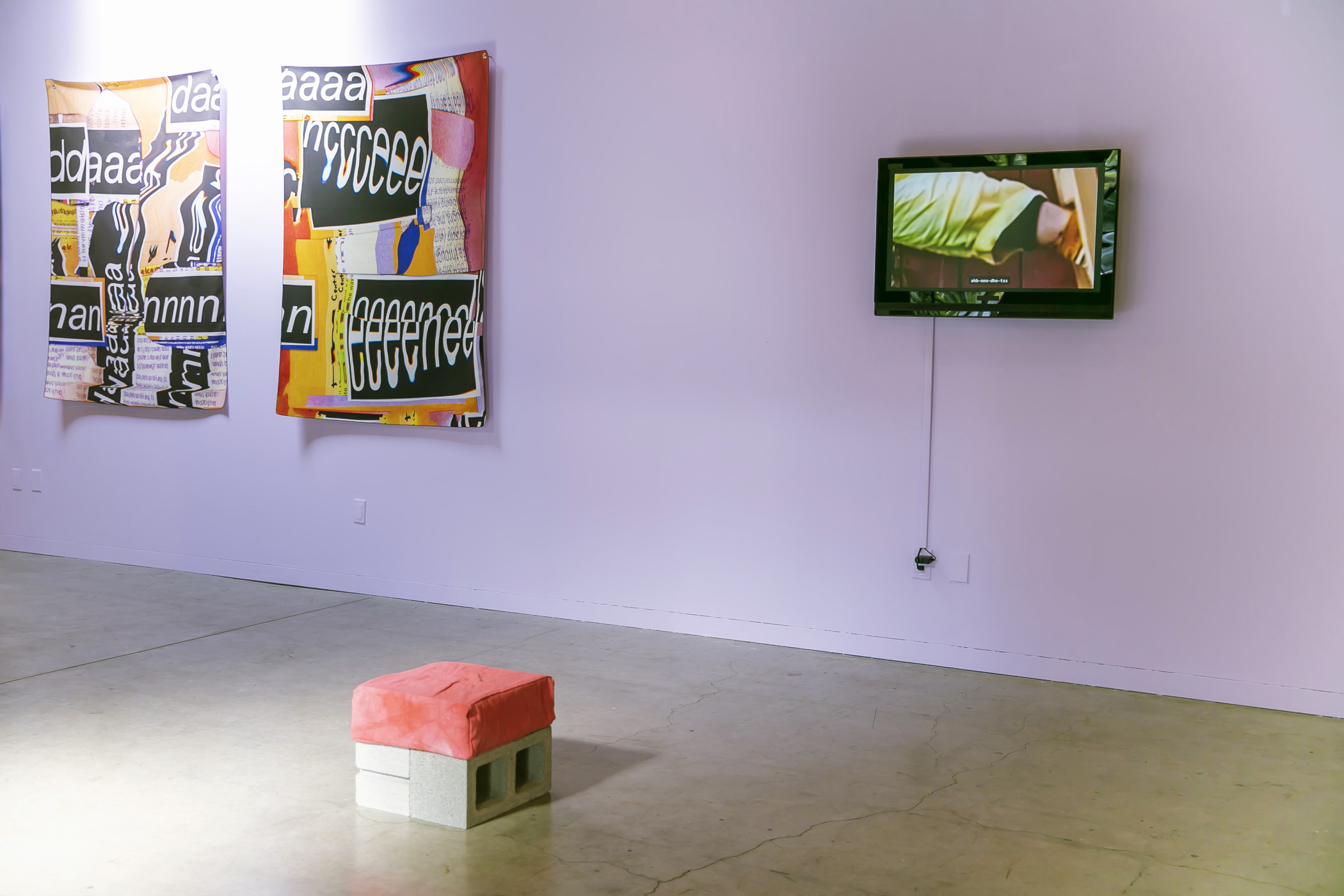 A purple wall with a screen. In front of the wall and on the floor sits two concrete blocks pushed together to make a seat. On top of them is a pink square, pink cushion. On the wall, to the left of the screen, is a wall hanging that is orange, black, and red. The screen shows a foot with the edge of a yellow pant, pushing up against something. 
