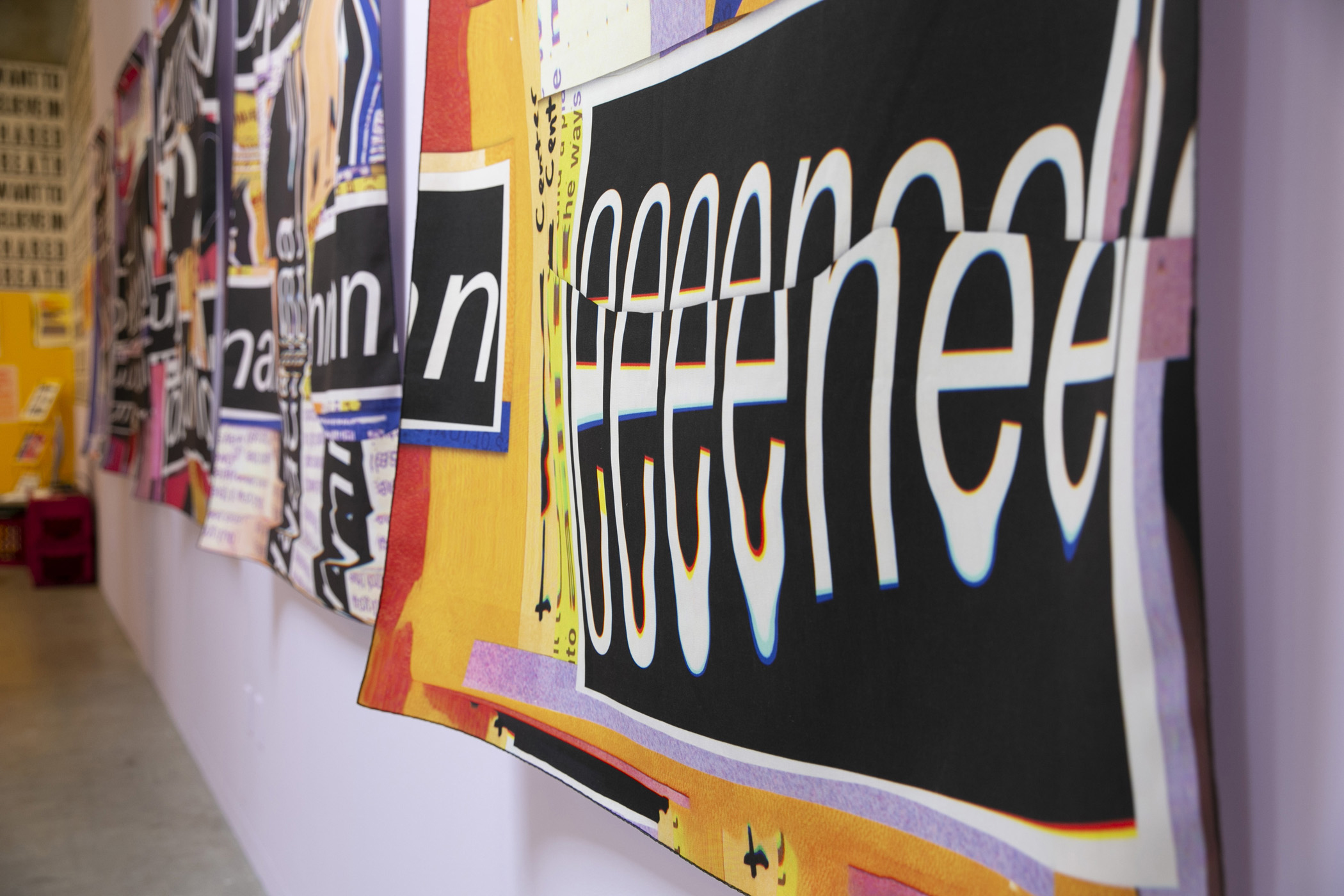 a close up of the wall hangings. We see a fabric hanging off of the purple wall and a close up of a string of ‘E’’s in white warped text that sit over a block of black. The banner hangs loosely, not very constrained to the wall. 
