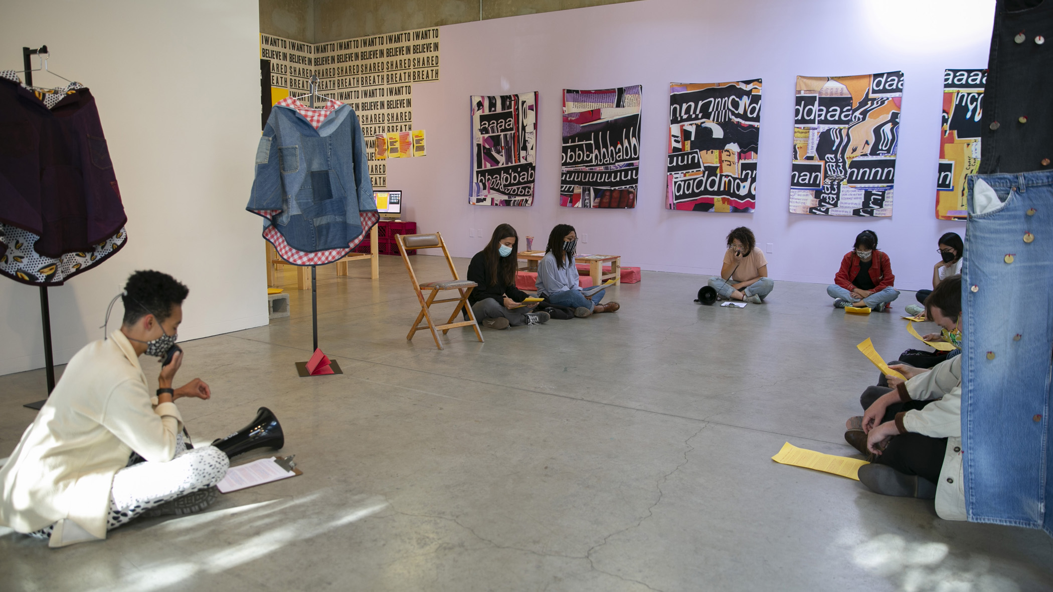 A group of people sit on the floor in a gallery. Two readers face each other at the edges of the circle, each holding the speaker component of a megaphone and looking downward at a text. They read this text together. Others sitting around in the circle are holding yellow flyers with the text, so they can read and speak along. 