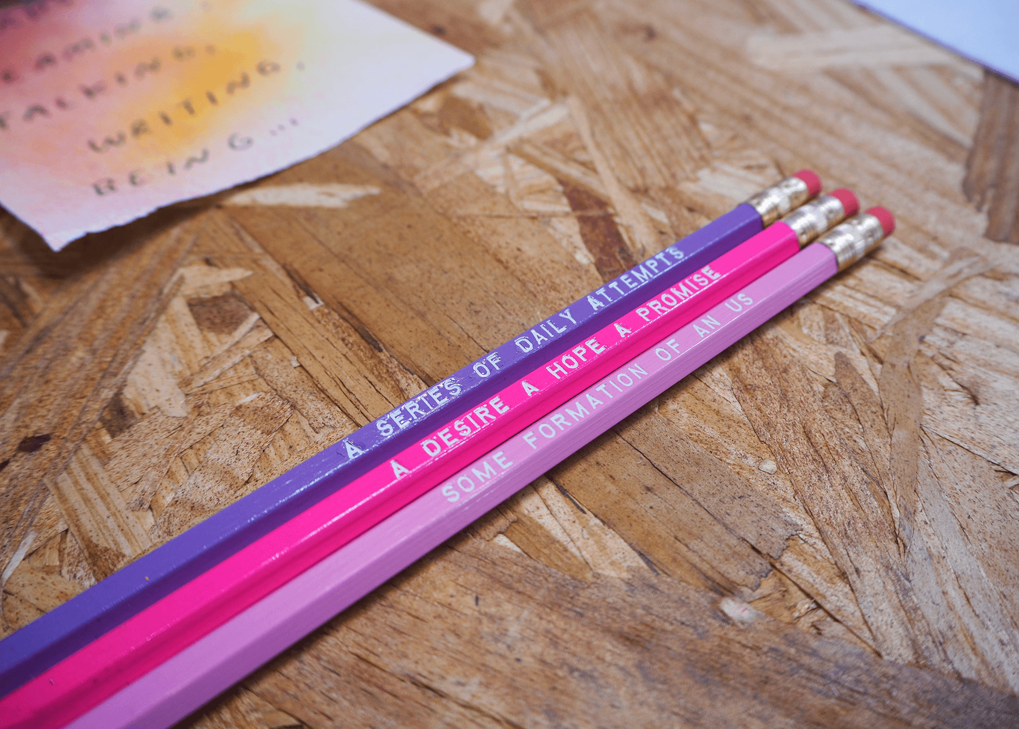 three pencils in lavender, bright pink, and light pink sit together on top of a wooden / OSB board background. In white text, each pencil has a line together reads, “a series of daily attempts; a desire a hope a promise; some formation of an us.” 

