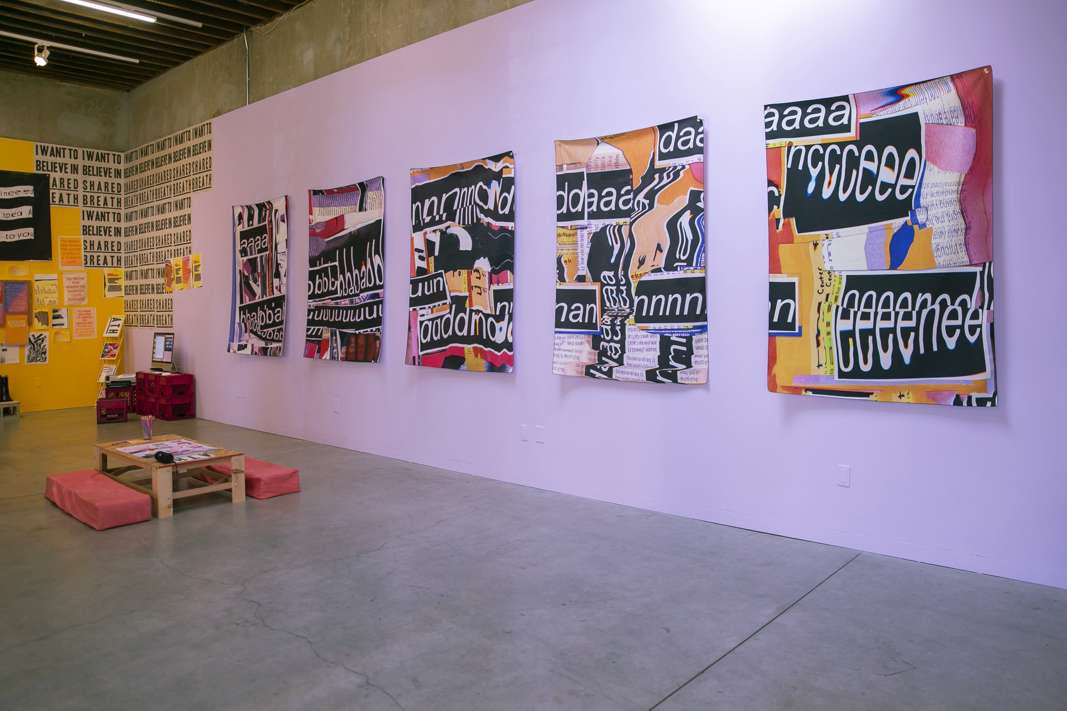 A purple wall is seen at an angle with five wall hangings on top of it. The wall hangings are made of fabric and have a collage feel. They are full of repeated letters that have been warped. The letters are white and have black backgrounds. They feel like cut pieces of paper that sit onto colorful collages. When read together, the banners spell out “abundance.” 
