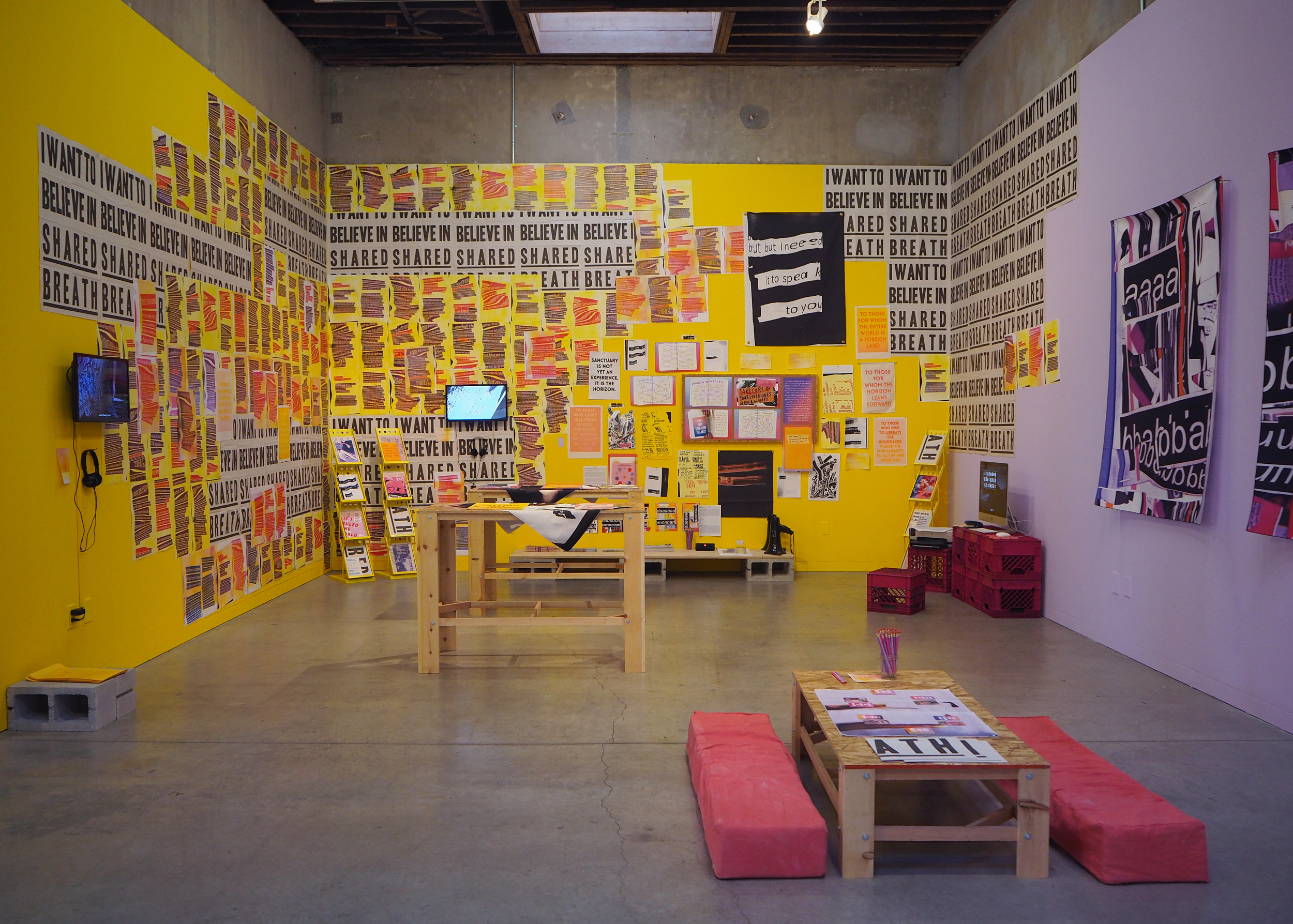 Another overview image of the same space but from a more central angle. The left and center walls are yellow and the right wall is purple. The yellow walls are covered in pink, purple, and grey prints. One dominant print reads, “I want to believe in shared breath” and is covered up and then revealed by different prints that have been patterned on top. The purple wall is more bare, with a small computer set up on the right.