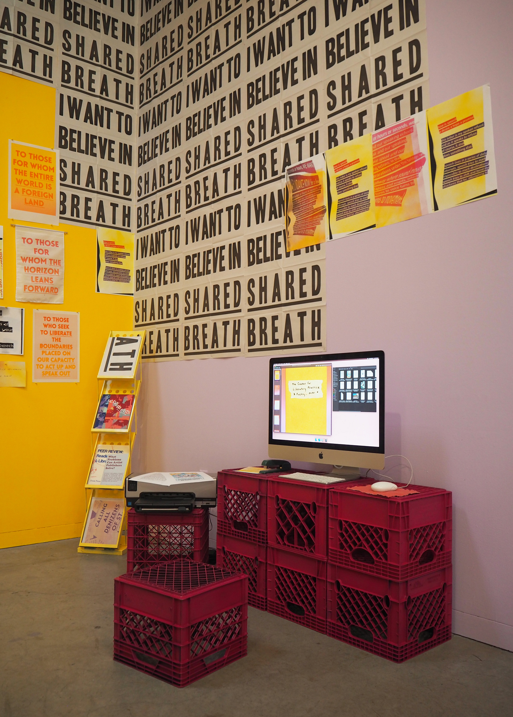 A computer sits on top of six redd-pinkish crates. To the left of the crates is a metallic, yellow publication stand. It has four levels and the structure feels like an accordion. Behind the computer is a purple wall that has some grey and black posters over top. The posters have bold text that says, “I want to believe in shared breath” in all capital letters. There are four smaller yellow posters with pink and purple blocks of text on top of the grey posters. They are on the wall in a horizontal line, slightly to the right of the computer. The most left wall, which we only see a fragment of, is yellow and has a few other printed pieces of text on it. 
