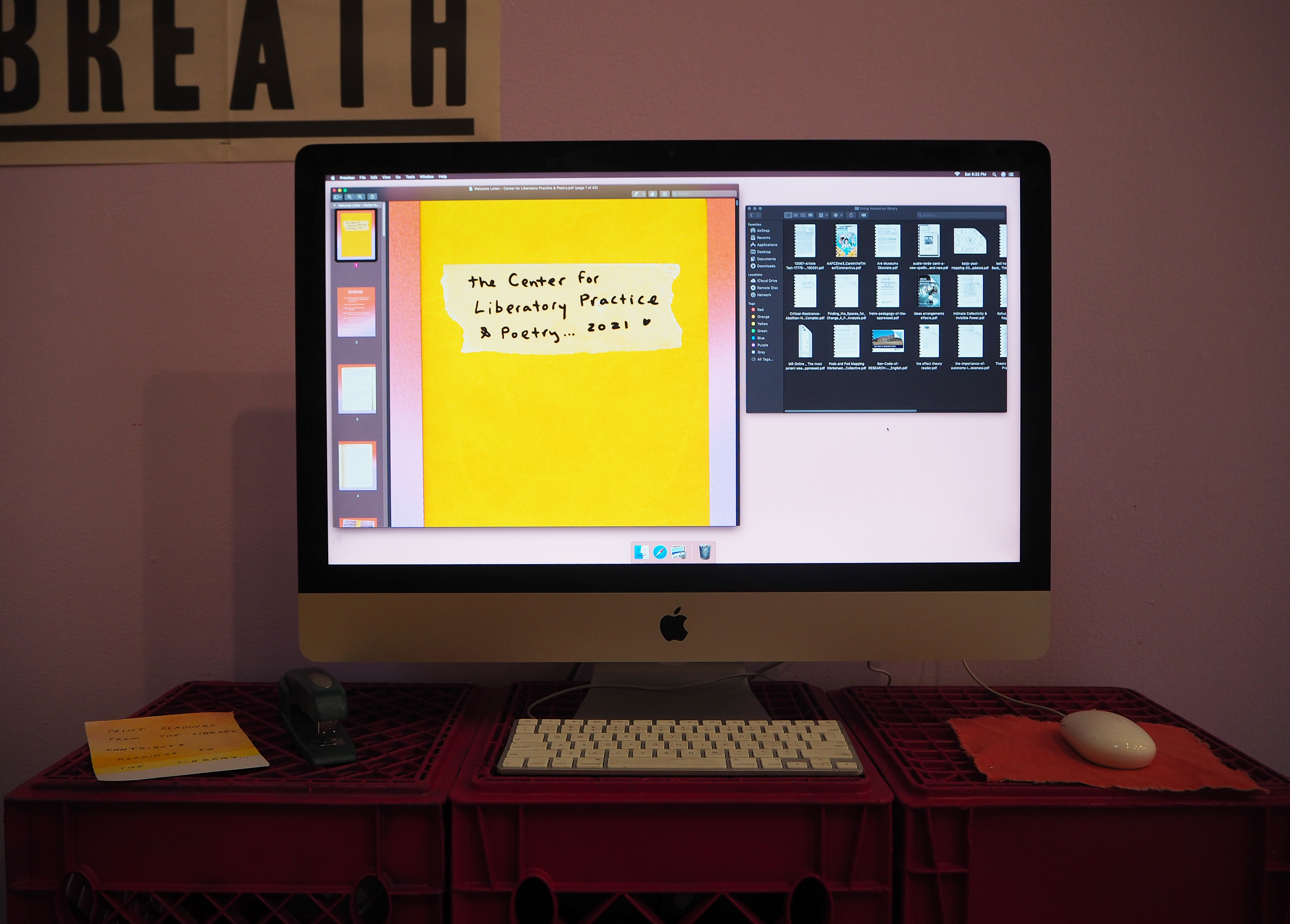 A computer screen is large and at the center of this image. It is sitting on three red-pink crates. The brightness of the screen makes the surroundings feel darker. Shades of purple and pink surround the screen. On the screen, there is an open folder with PDFs inside and one document open. The document is open to a yellow page with small hand written text on top that reads, “The Center for Liberatory Practice & Poetry….2021 <3”. 
