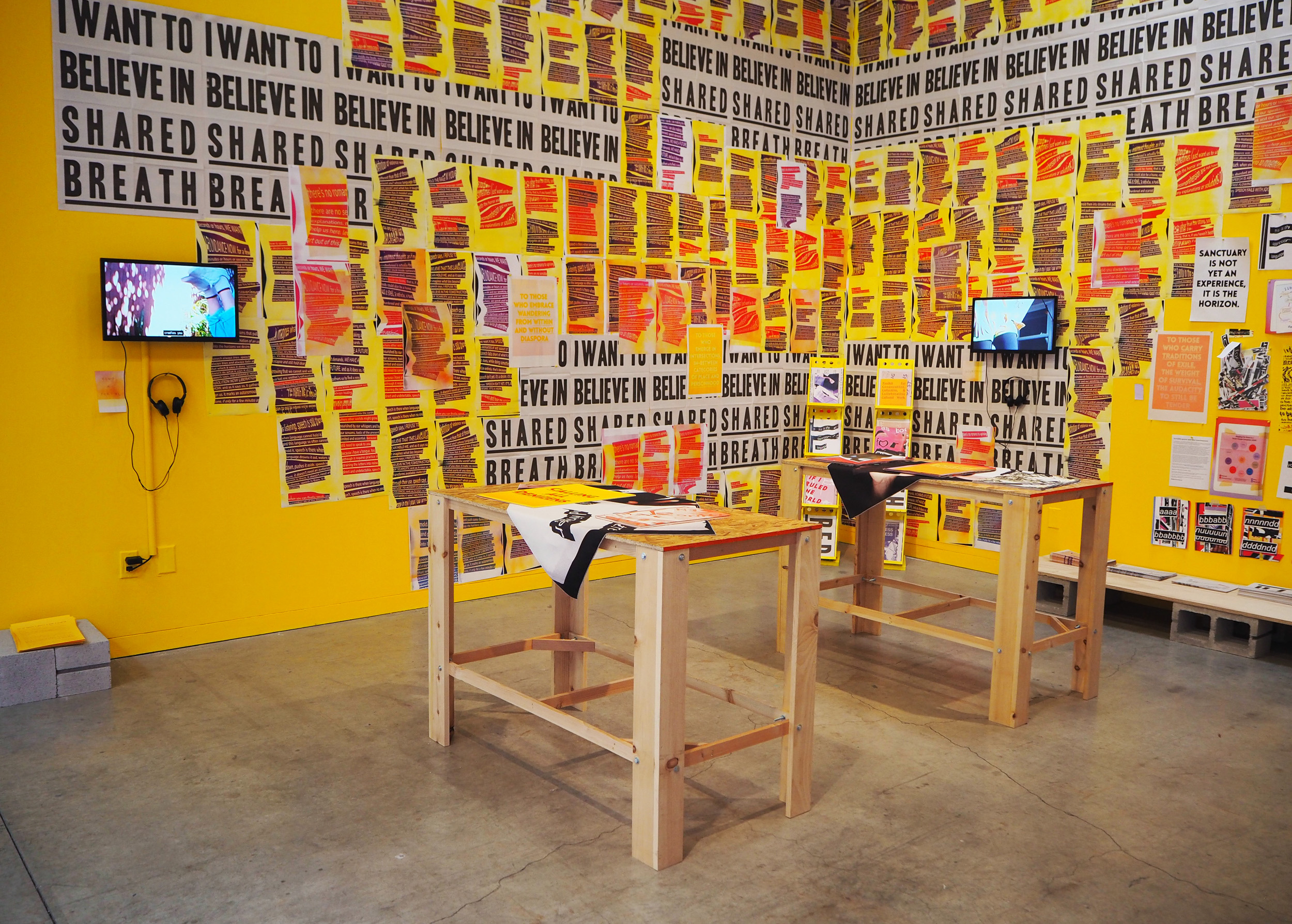 An overview of the main space that centers on two yellow walls covered in posters. Two work tables stand in the middle, they are made of wood and have fabric draping over them that doesn’t cover the entire table top. The posters on the walls have pink and purple blocks of text over yellow and orange backgrounds. Other posters are grey with bold black text overtop that reads, “I want to believe in shared breath.” Two small screens are also hung above the posters. One screen shows the image of feet floating in the air, the other screen shows an image of legs in movement. The floor is grey and slightly reflects the light from the yellow walls. 
