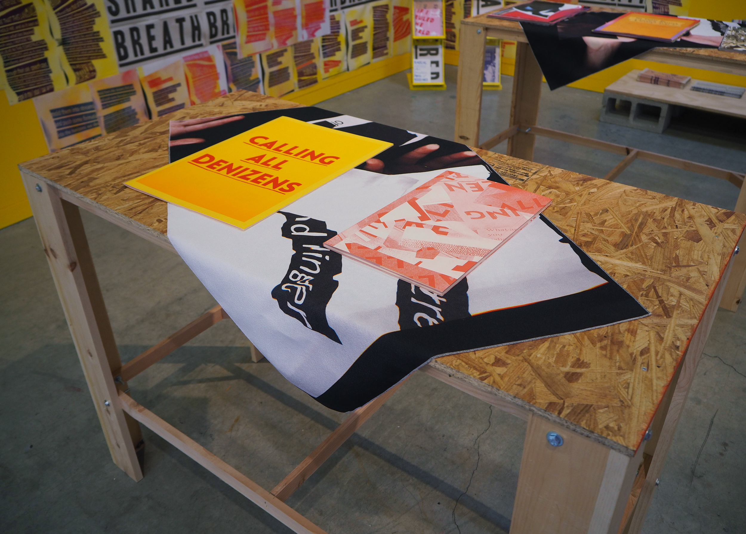 A closer look at two books laying on a table. The table top is made of OSB with a fabric draping over its center diagonally. The left and right sides of the tabletop are revealed. The fabric is white and black and feels like a xerox print of warped lettering. The most left book is a large yellow zine with reddish bold text that reads, “Calling all Denizens”. The other book has a red and white, abstract collage. 