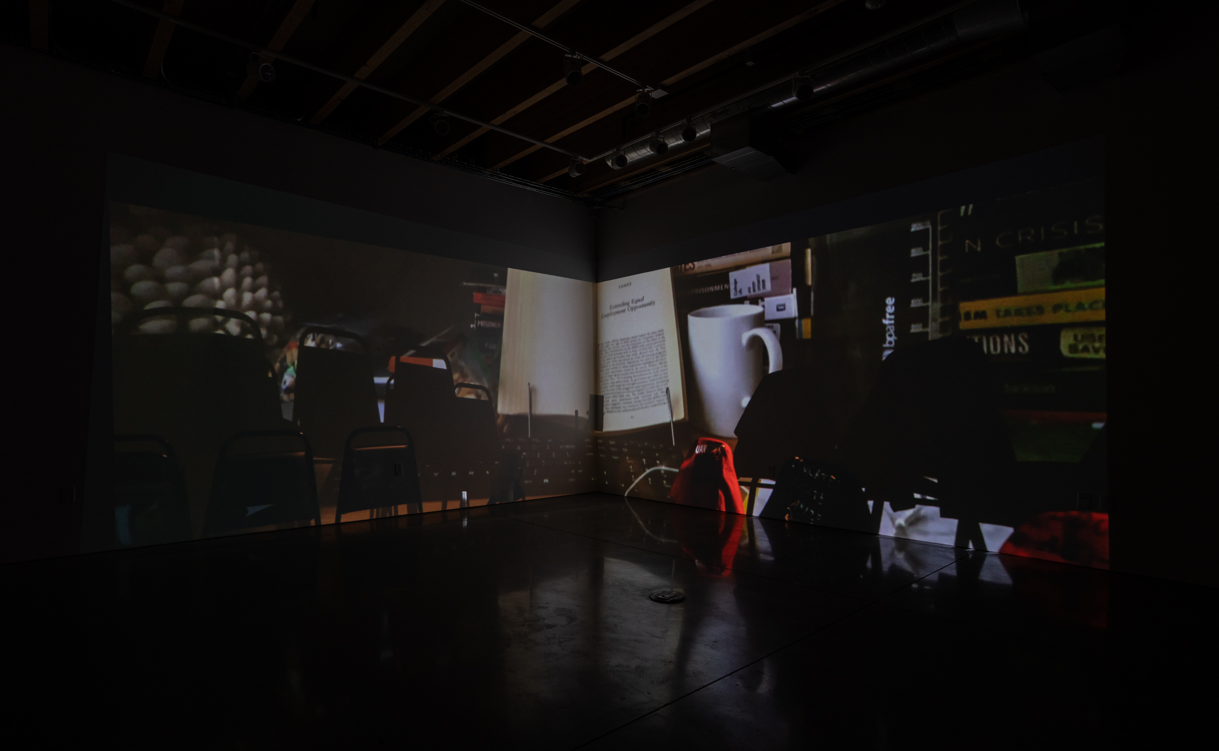 Installation shot of Stage Directions (UAW 2865) a two-channel video projected into the corner of a room. The image is dark but the light from an overhead projector in a rhombus shape taking up the center of the image on both screens. I n large, bold, white text “Stage” is written in the upper left corner. In the bottom right “Directions”.
