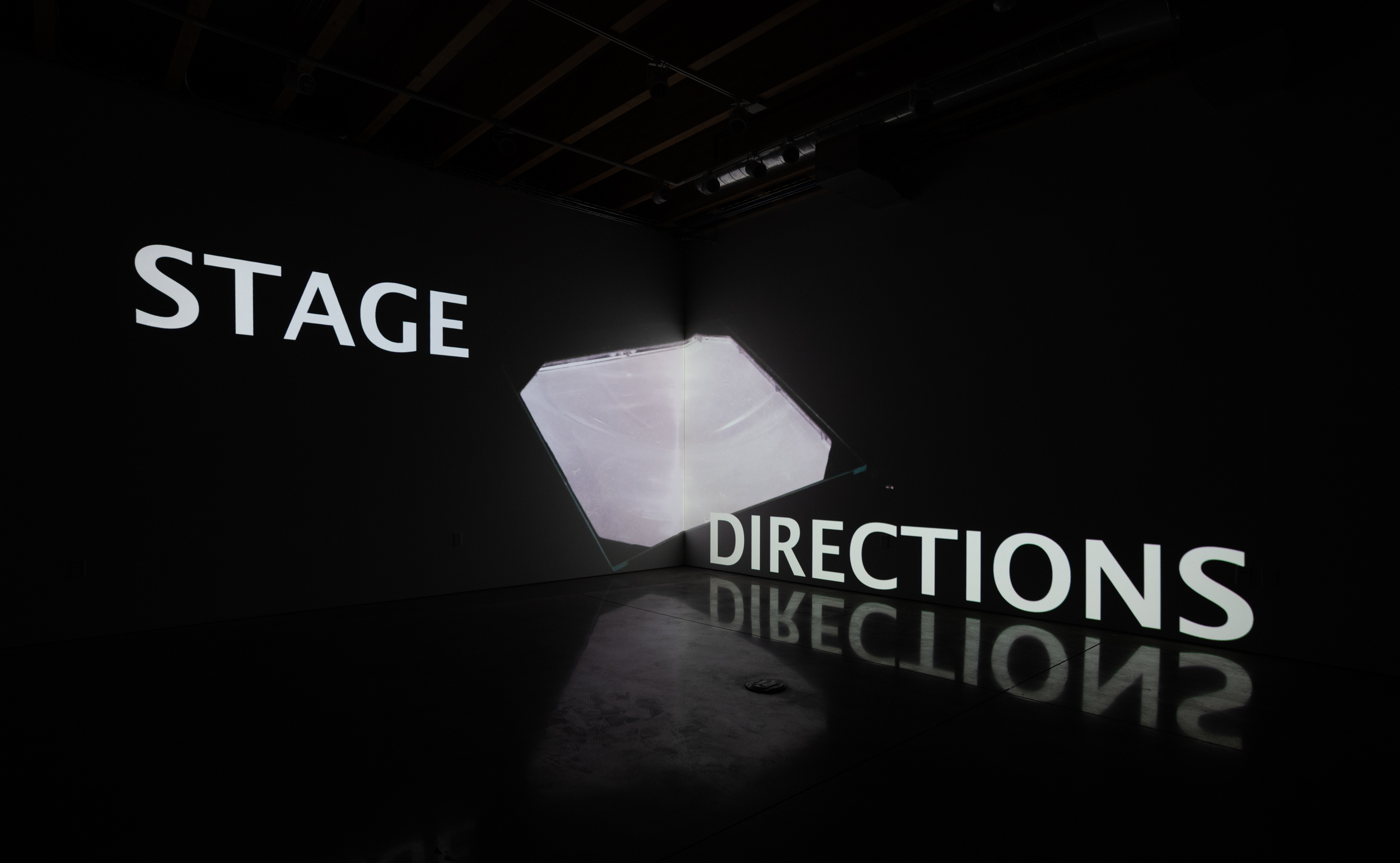  Installation shot of Stage Directions (UAW 2865) a two-channel video projected into the corner of a room. Across both screens is an image of members of a theater group with their bodies entangled as they take part in a movement workshop. 
