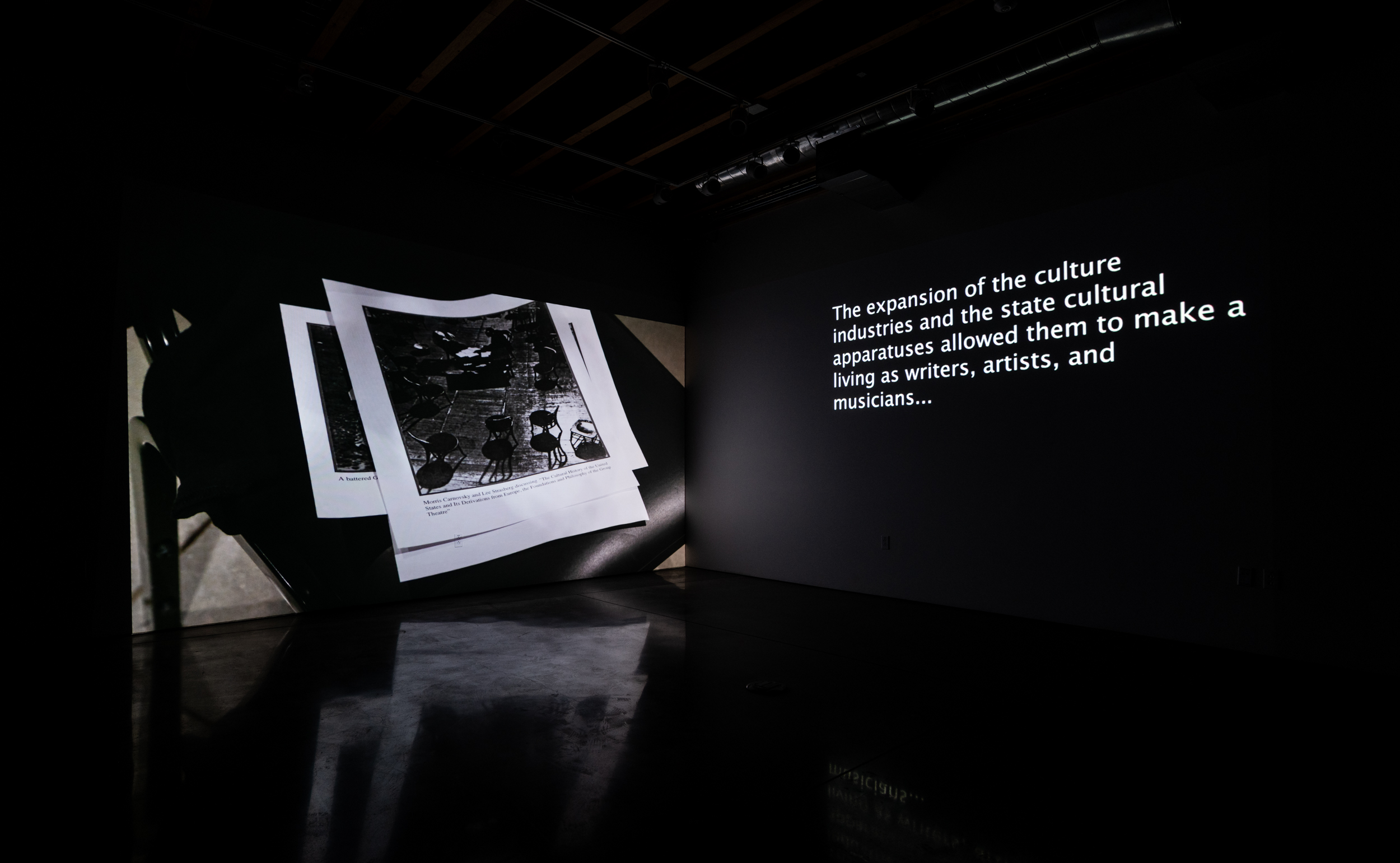 Installation shot of Stage Directions (UAW 2865) a two-channel video projected into the corner of a room. On the left screen is a close up of a desk. On it there is a computer monitor, a keyboard, a mouse and stationary surrounded by piles of papers. On the right screen is phone footage of a pile of UAW picket signs on the floor. 