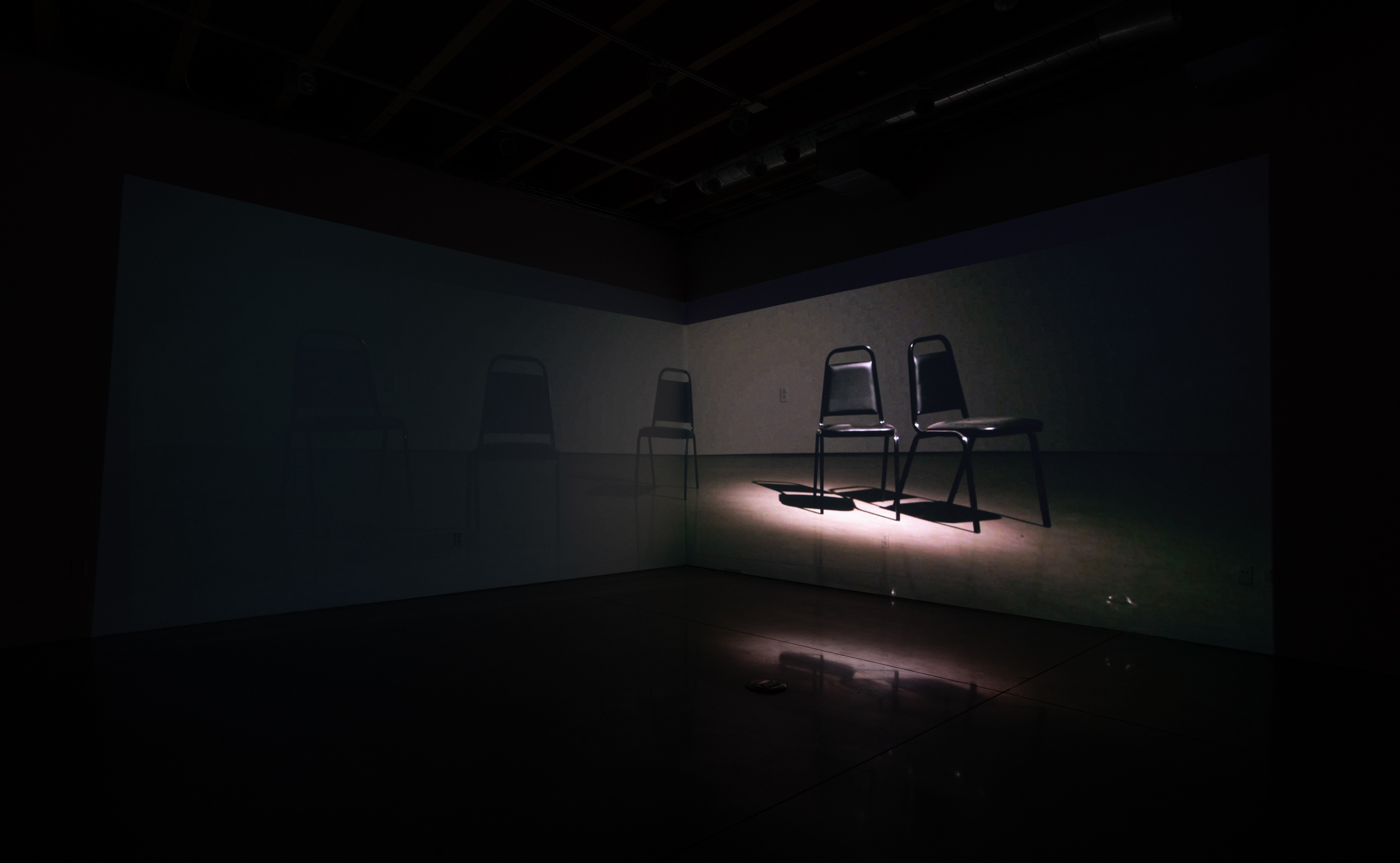 Installation shot of Stage Directions (UAW 2865) a two-channel video projected into the corner of a room. The left side of the image is dark, on the right there is a spotlight on two chairs angled slightly away from one another and facing towards the audience. In the center is another chair slightly lit up. 
