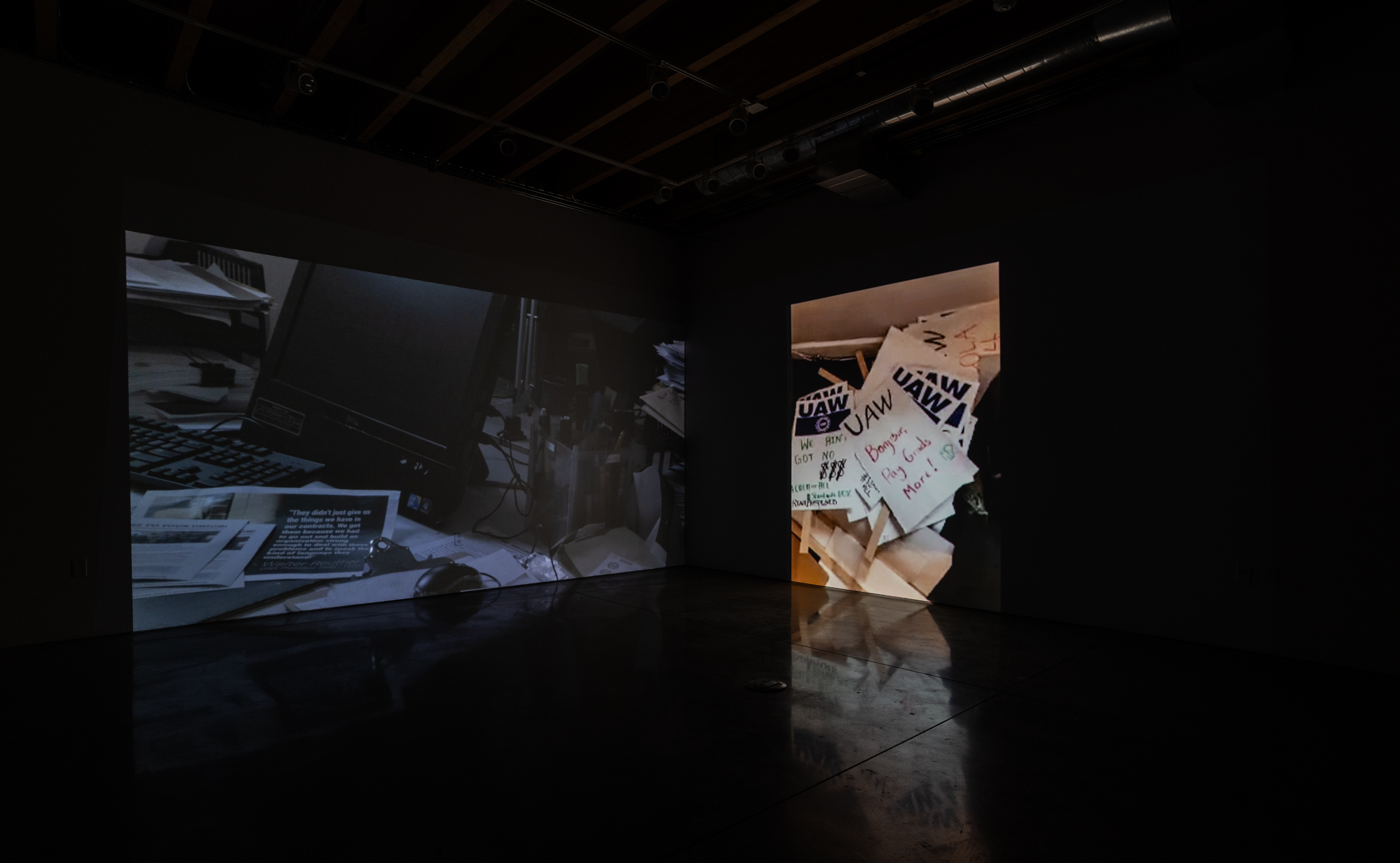 Installation shot of Stage Directions (UAW 2865) a two-channel video projected into the corner of a room. On the left screen is a close up of a pile of paper on a chair. The top sheet has an image with the caption “Morris Carnovsky and Lee Strasberg discussing  “The Cultural History of the United States and Its Derivations from Europe, the Foundations and Philosophy of the Group Theatre”. On the right screen there is white text on a black background. It reads “The expansion of the culture industries and the state cultural apparatuses allowed them to make a living as writers, artists and musicians…”
