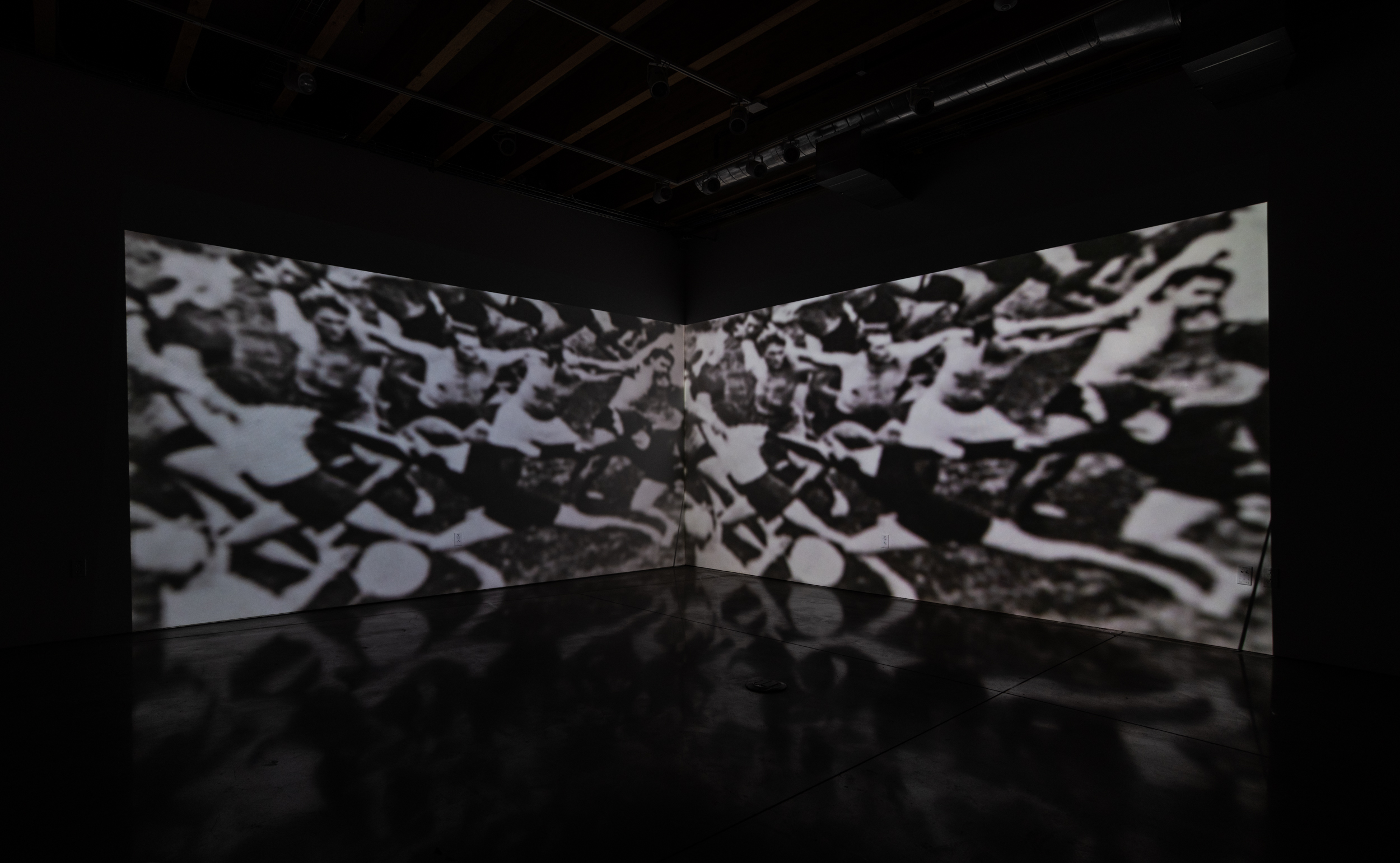Installation shot of Stage Directions (UAW 2865) a two-channel video projected into the corner of a room. In the center of the image is a book open to “Chapter 3: Expanding Equal Employment Opportunity”. Next to that on the right a half drunk from coffee mug. Behind that are a pile of books, legible on the spines are the words “Crisis”, “Takes Place”, “Used Saves,” “Imprisonment”, “In the City”. Shadows of chairs can be seen in the foreground. 
